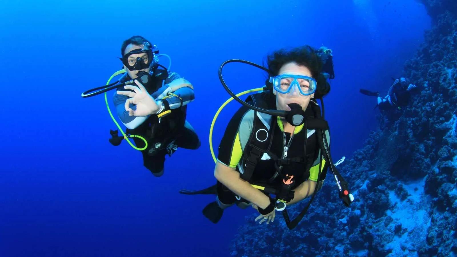 Two scuba divers giving the OK sign to camera, a third diver visible near the reef behind them