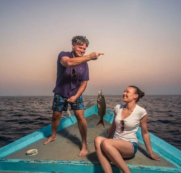 A guest holding up a freshly caught fish on a traditional dhoni at dusk, another guest looking on