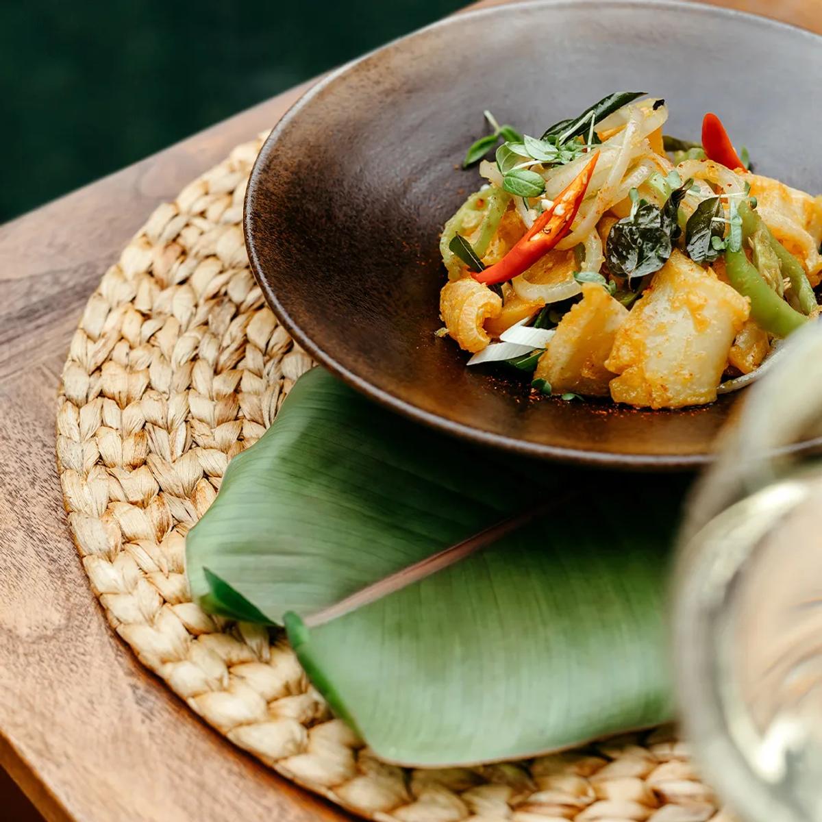 A plated Maldivian-style curry with banana leaf garnish and a glass of wine, representative of casual dining across Iru Fushi's poolside outlets — dedicated Fluid photography to follow