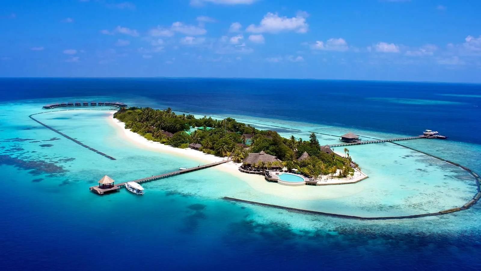 Aerial view of Komandoo island at midday, showing the freshwater infinity pool, main jetty and beachfront villas along the tree line