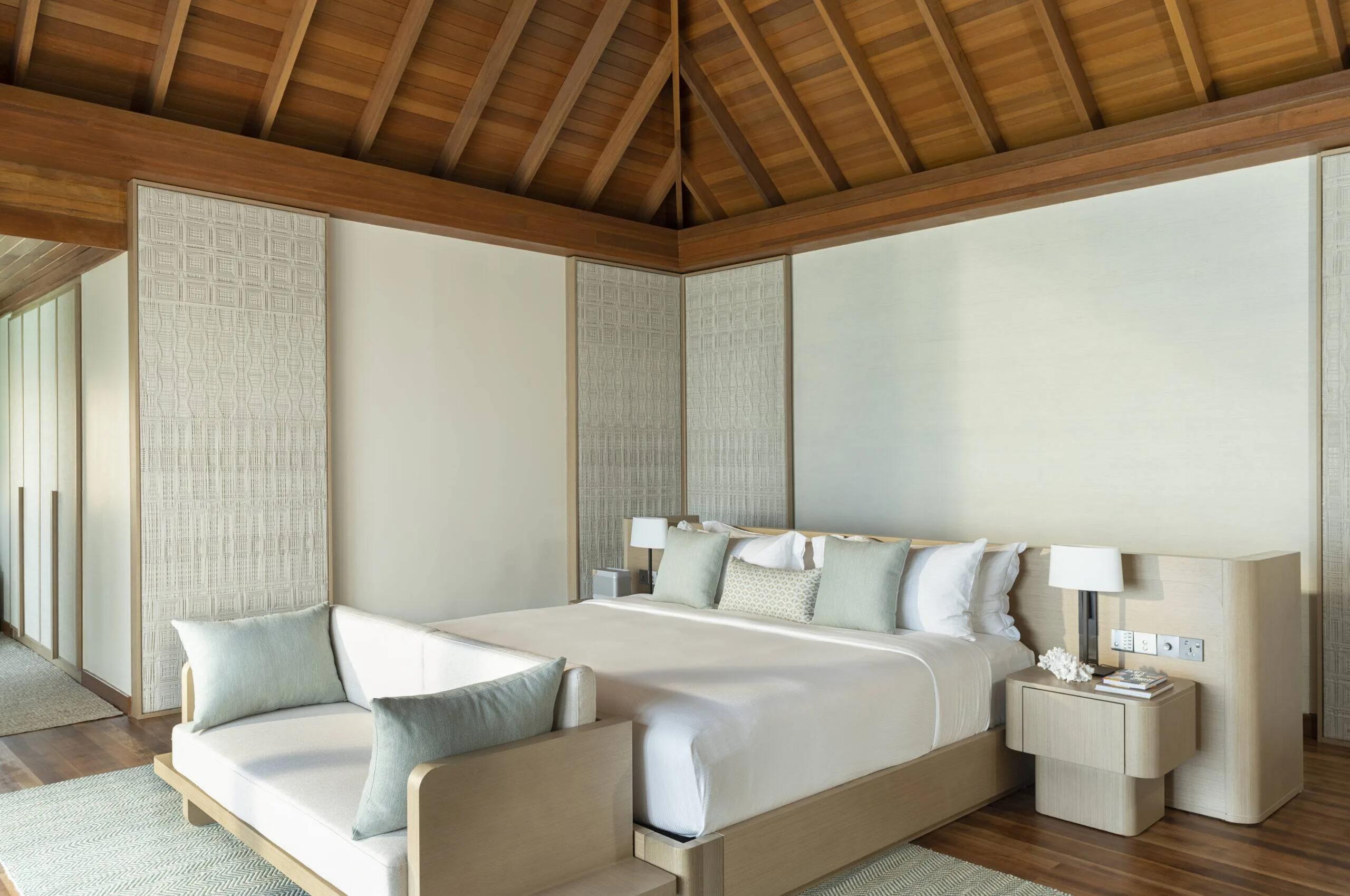 The bedroom of an Ocean Bungalow with Pool, a king bed dressed in teal and white beneath a peaked wood-raftered ceiling