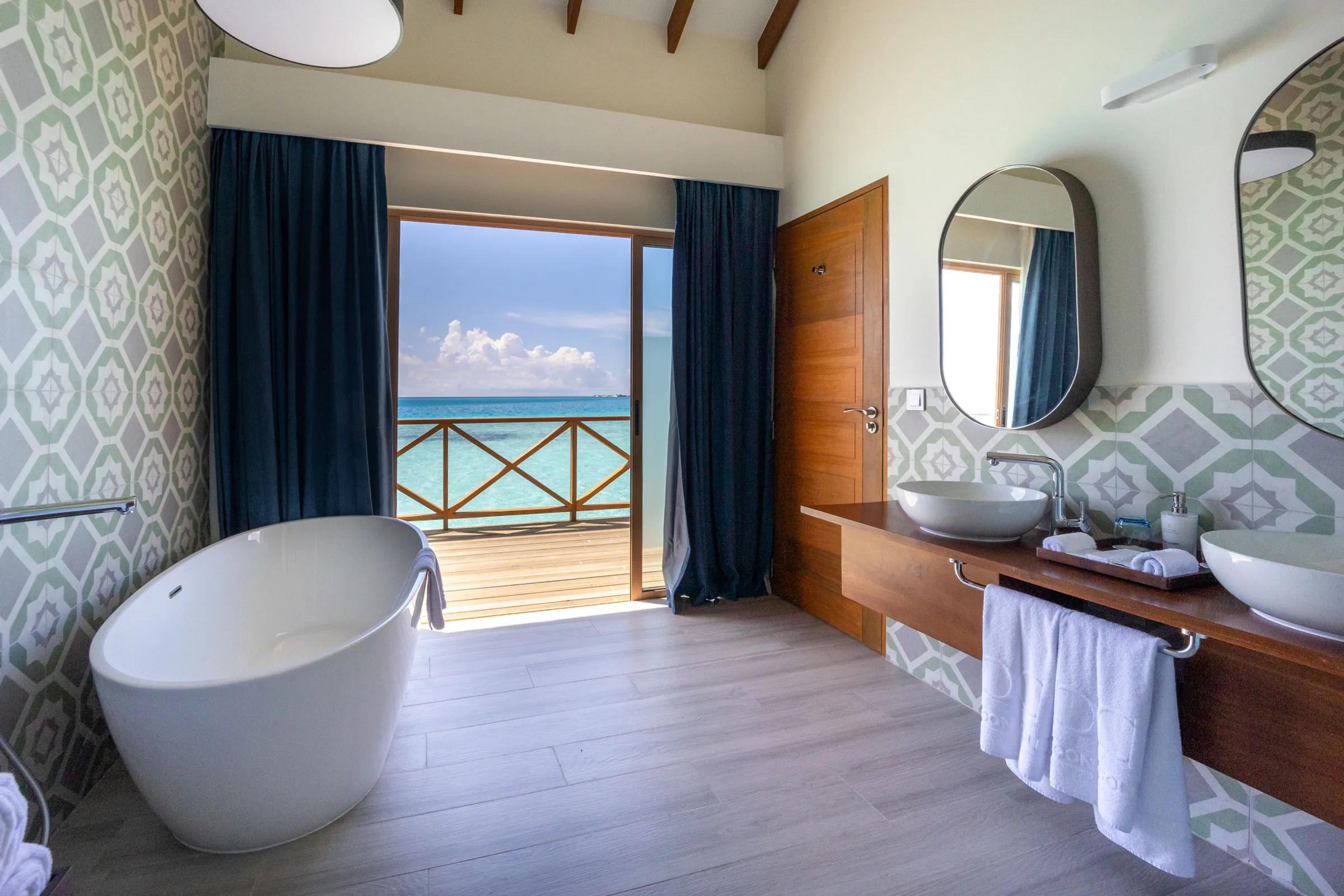 A Lagoon Suite's freestanding bathtub beside glass doors opening onto its overwater deck and the lagoon