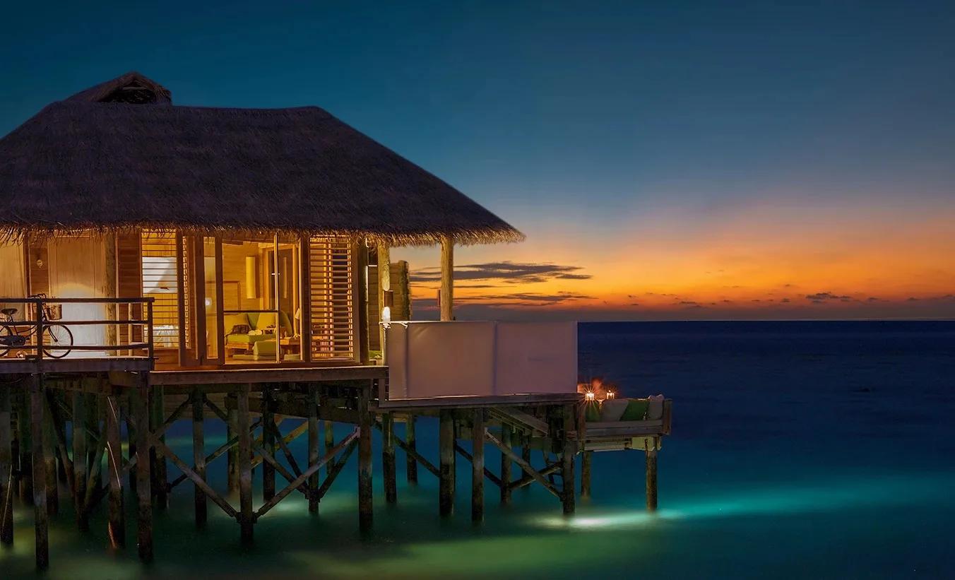 A water villa lit from within at dusk, its pool and deck facing an orange sunset horizon