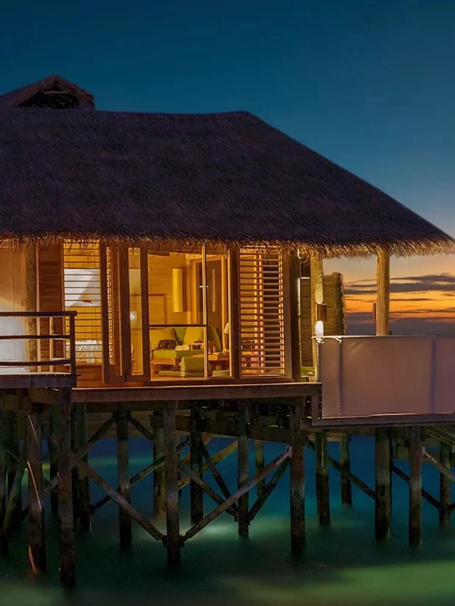 A water villa lit from within at dusk, its pool and deck facing an orange sunset horizon