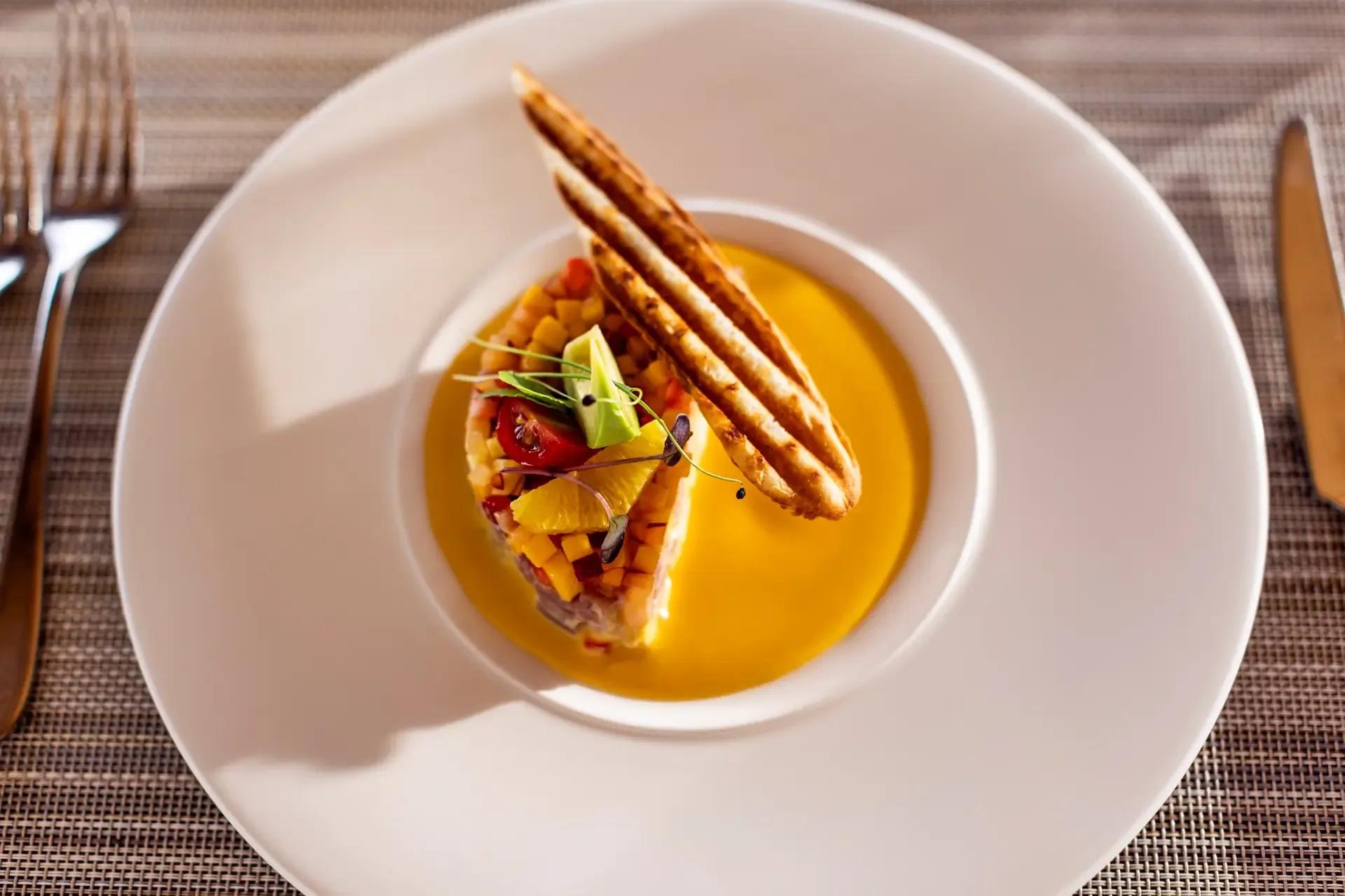 A layered seafood tartare with mango, tomato and a crisped bread wafer, set in a pool of citrus sauce