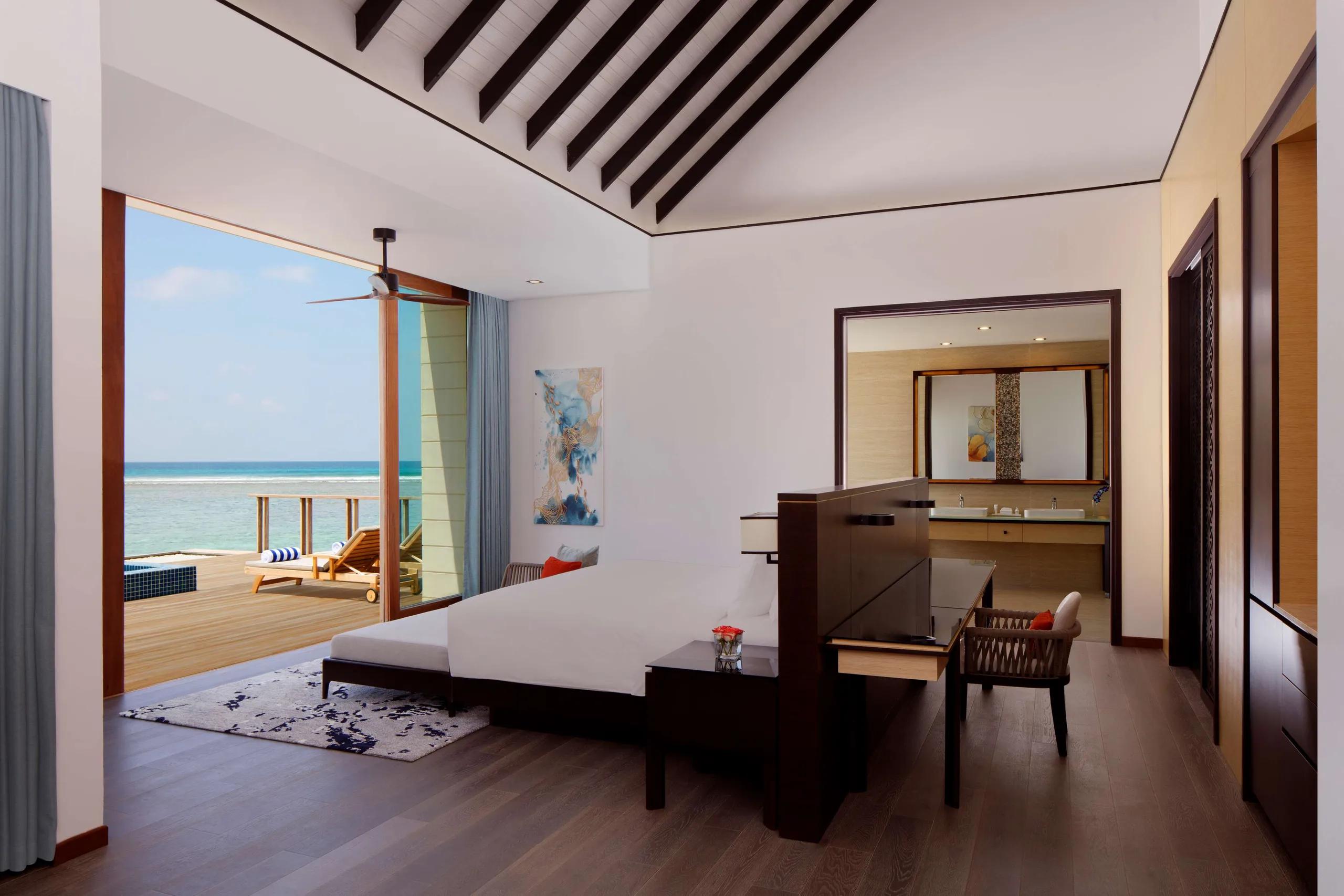 The bedroom of an Overwater Villa at Radisson Blu Resort Maldives, glass doors open to its private pool deck and the lagoon beyond