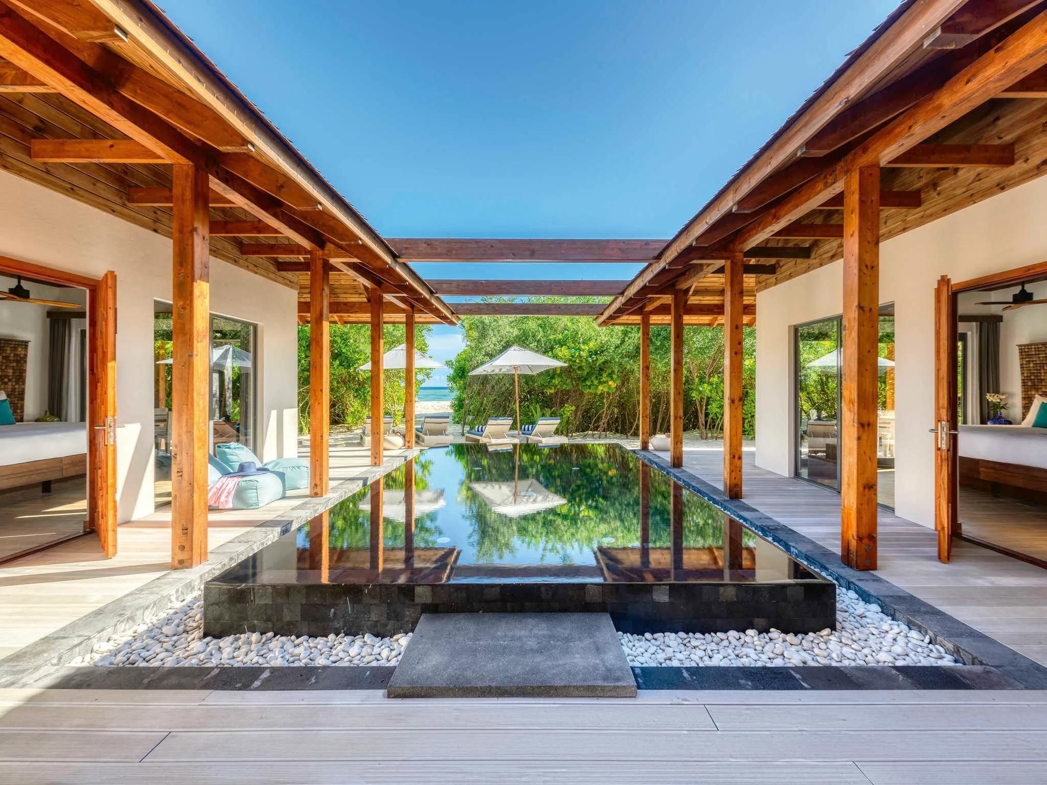 A long private pool running between two covered walkways connecting the bedroom pavilions of a beach residence, palm trees and beach visible at the far end