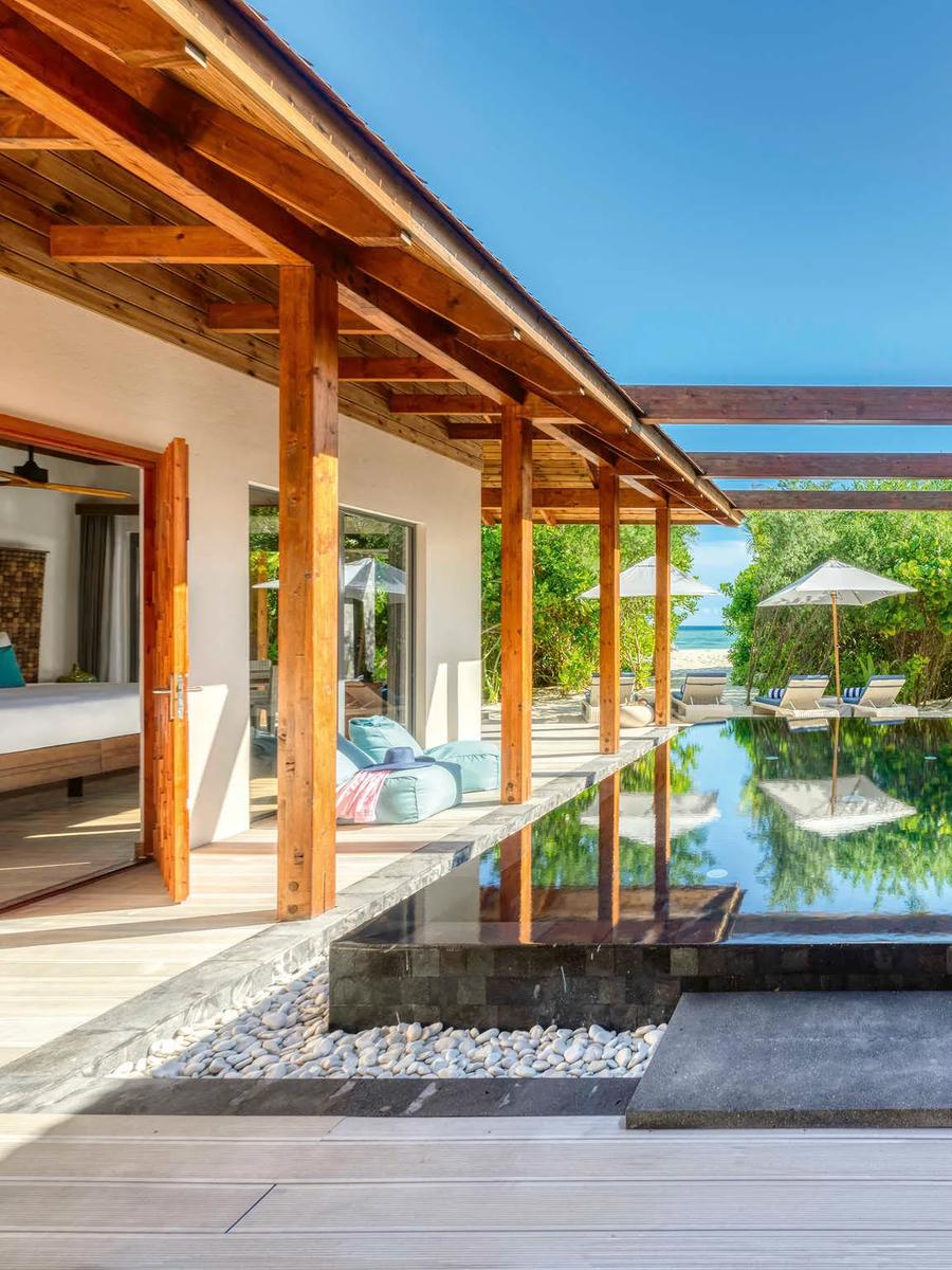 A long private pool running between two covered walkways connecting the bedroom pavilions of a beach residence, palm trees and beach visible at the far end