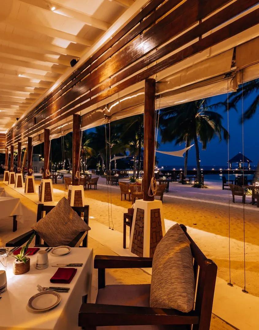 Sangu Beach's open-air dining room lit for the evening, tables looking out toward the beach and sea
