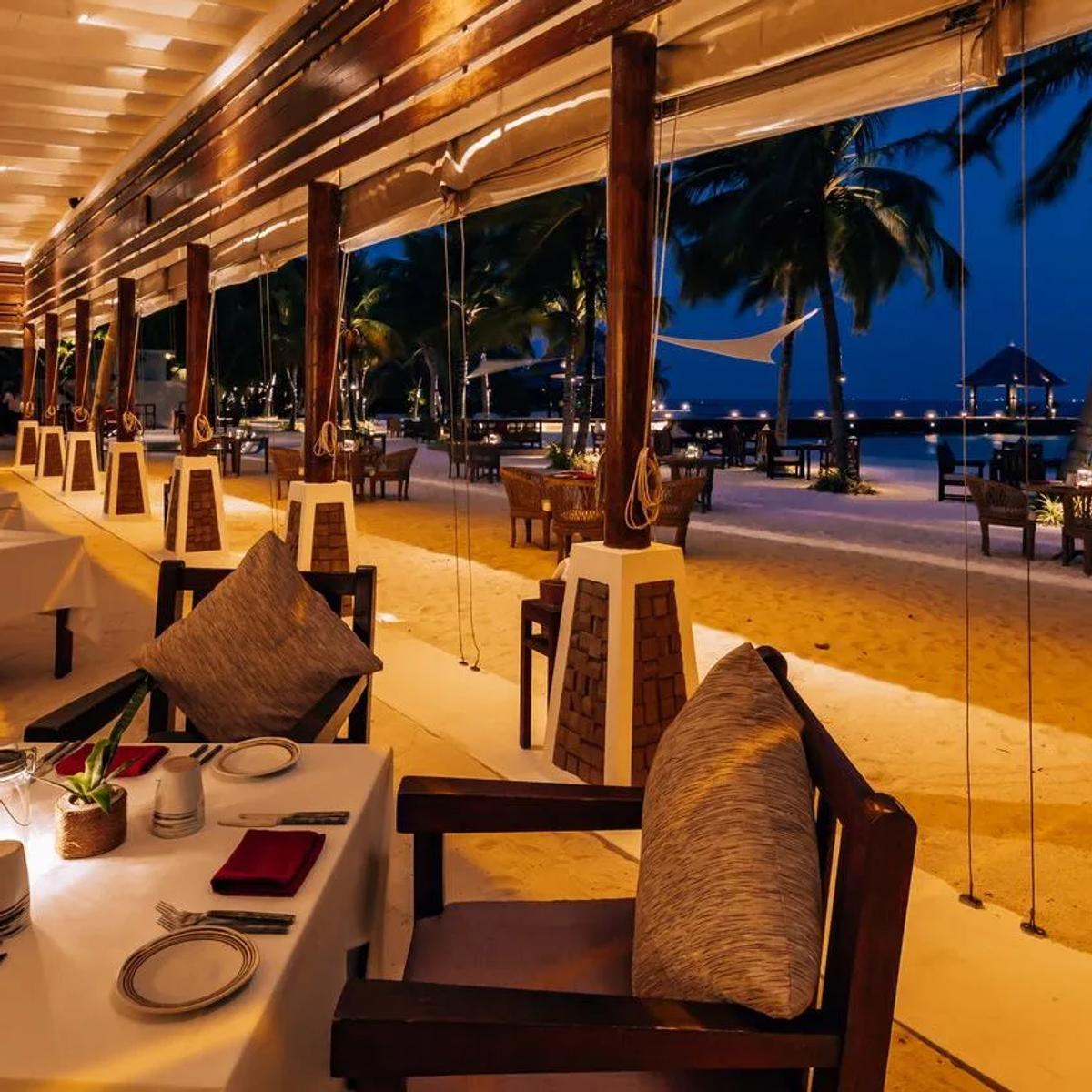 Sangu Beach's open-air dining room lit for the evening, tables looking out toward the beach and sea