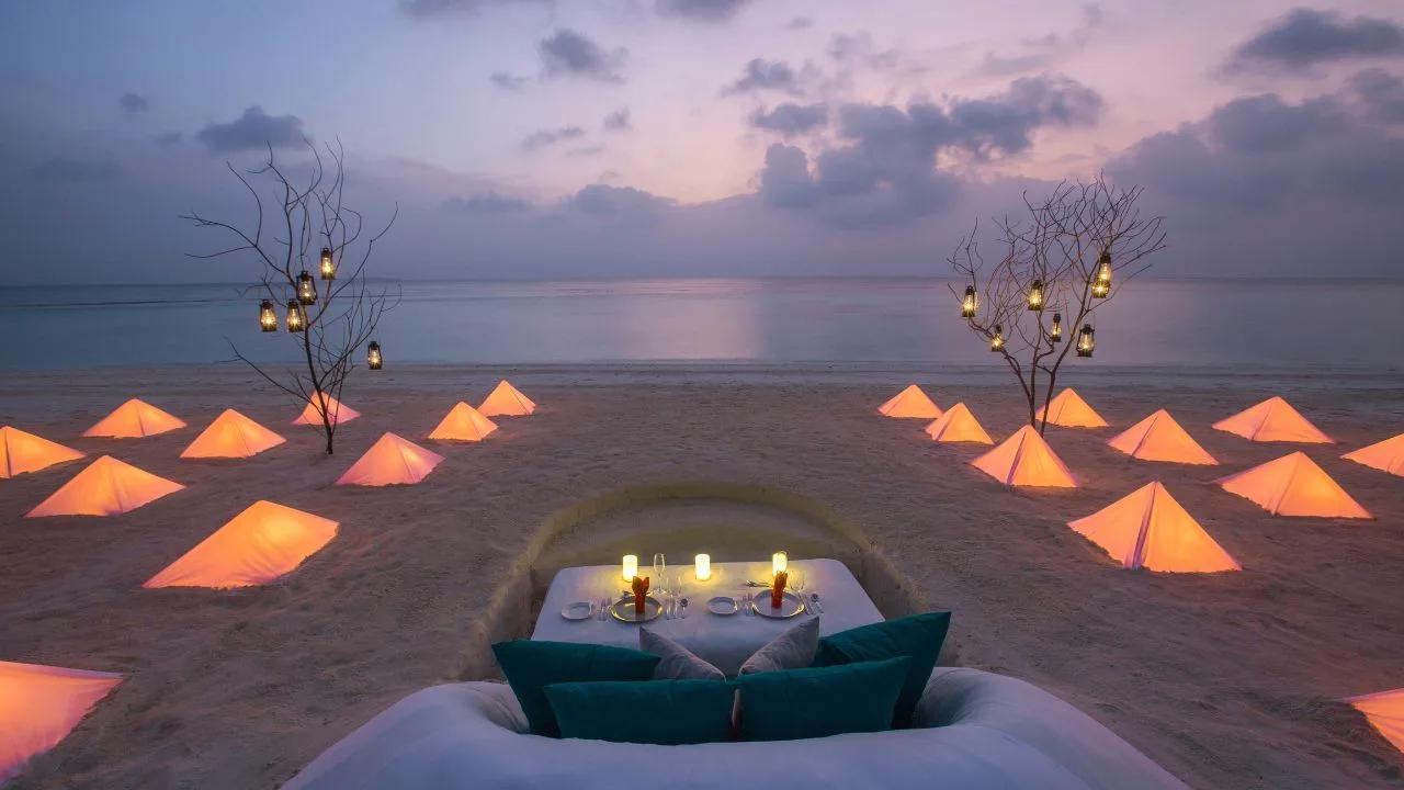 An illuminated private dining table set up on the beach at dusk, lanterns hung from bare branches on either side — Dusit Thani Maldives' Borderless Dining concept