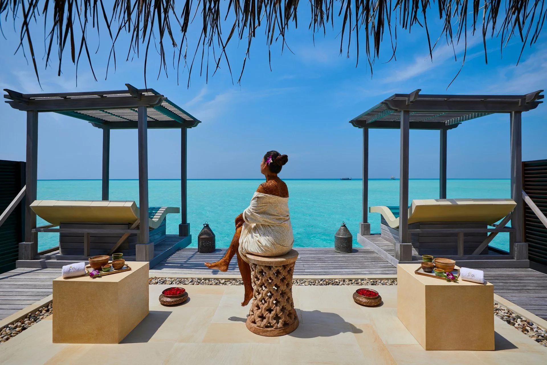 A guest wrapped in cloth after a mud or herbal body treatment, seated on an outdoor stool between two J Wellness Circle pergolas over the lagoon