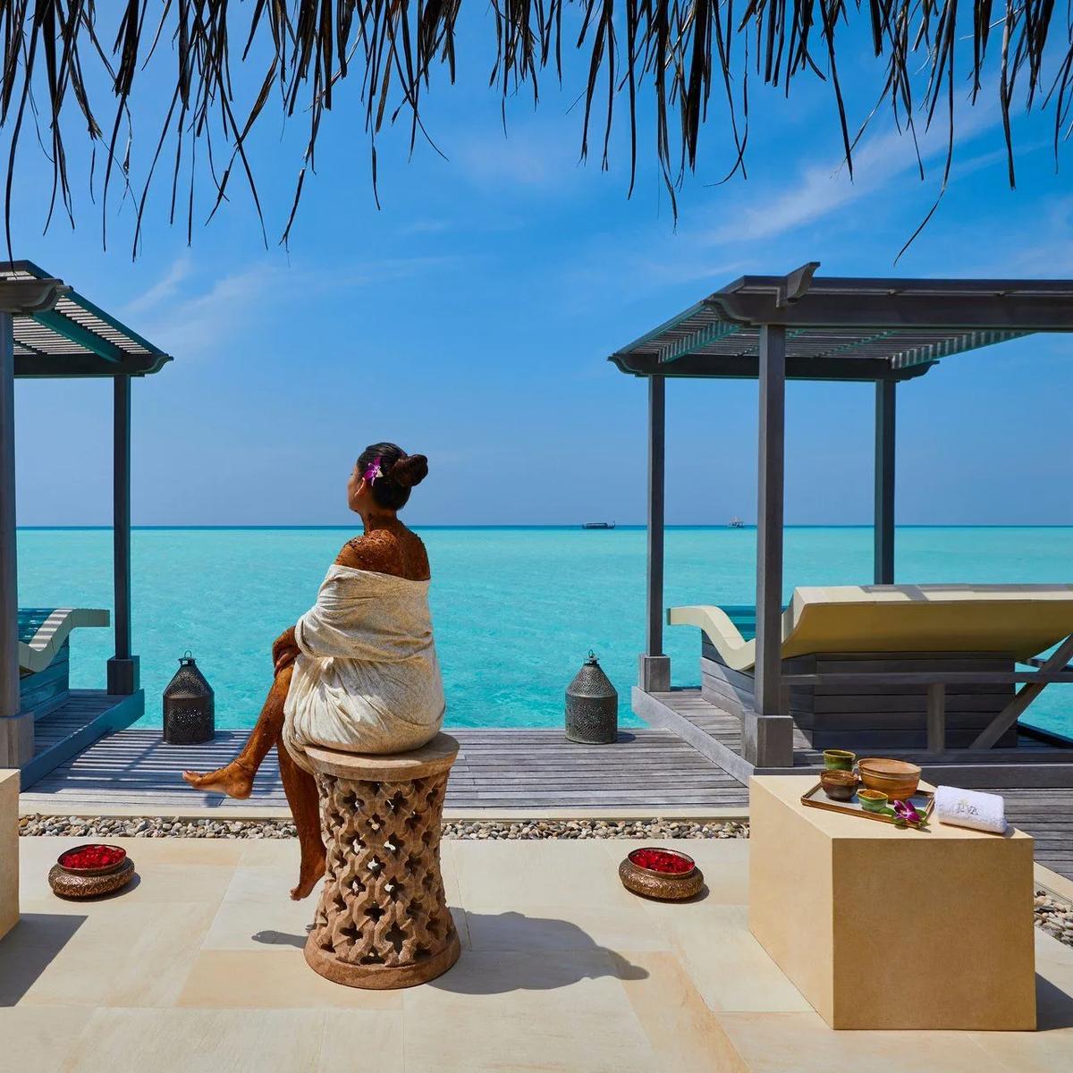 A guest wrapped in cloth after a mud or herbal body treatment, seated on an outdoor stool between two J Wellness Circle pergolas over the lagoon