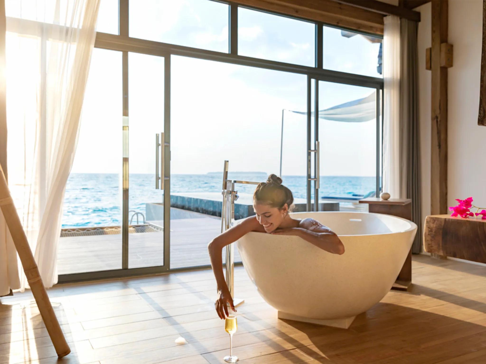 A woman relaxing in a freestanding bathtub beside floor-to-ceiling windows open to the ocean, a glass of champagne on the floor nearby