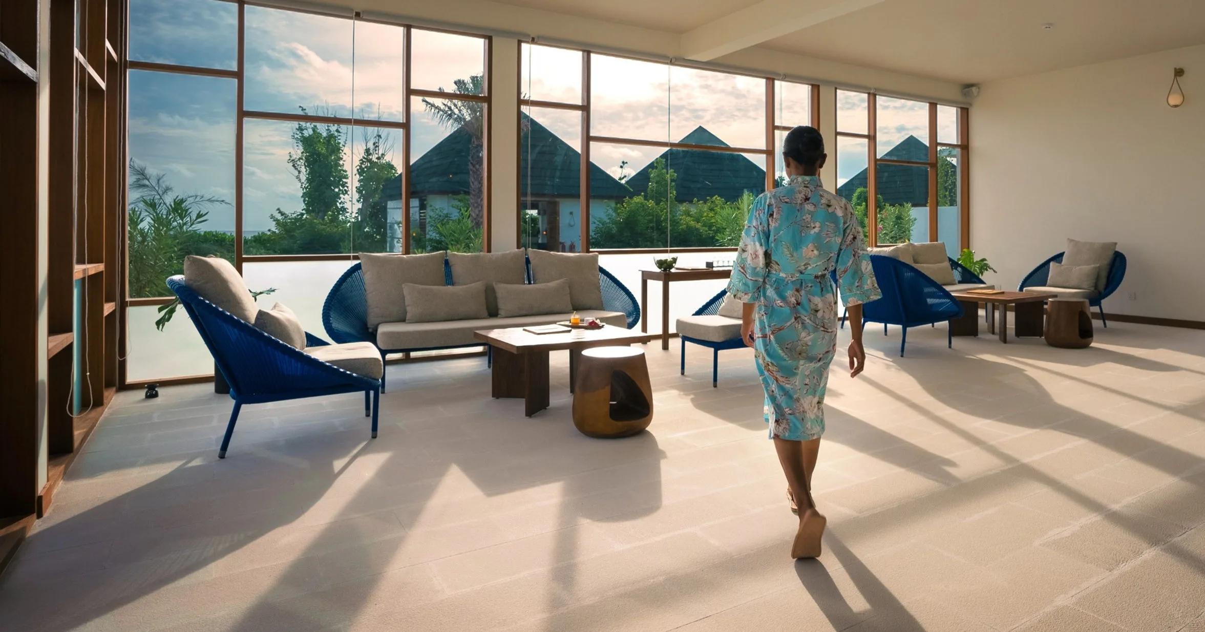 A guest in a robe walks through Oasis Spa's relaxation lounge, thatched treatment pavilions visible through the windows