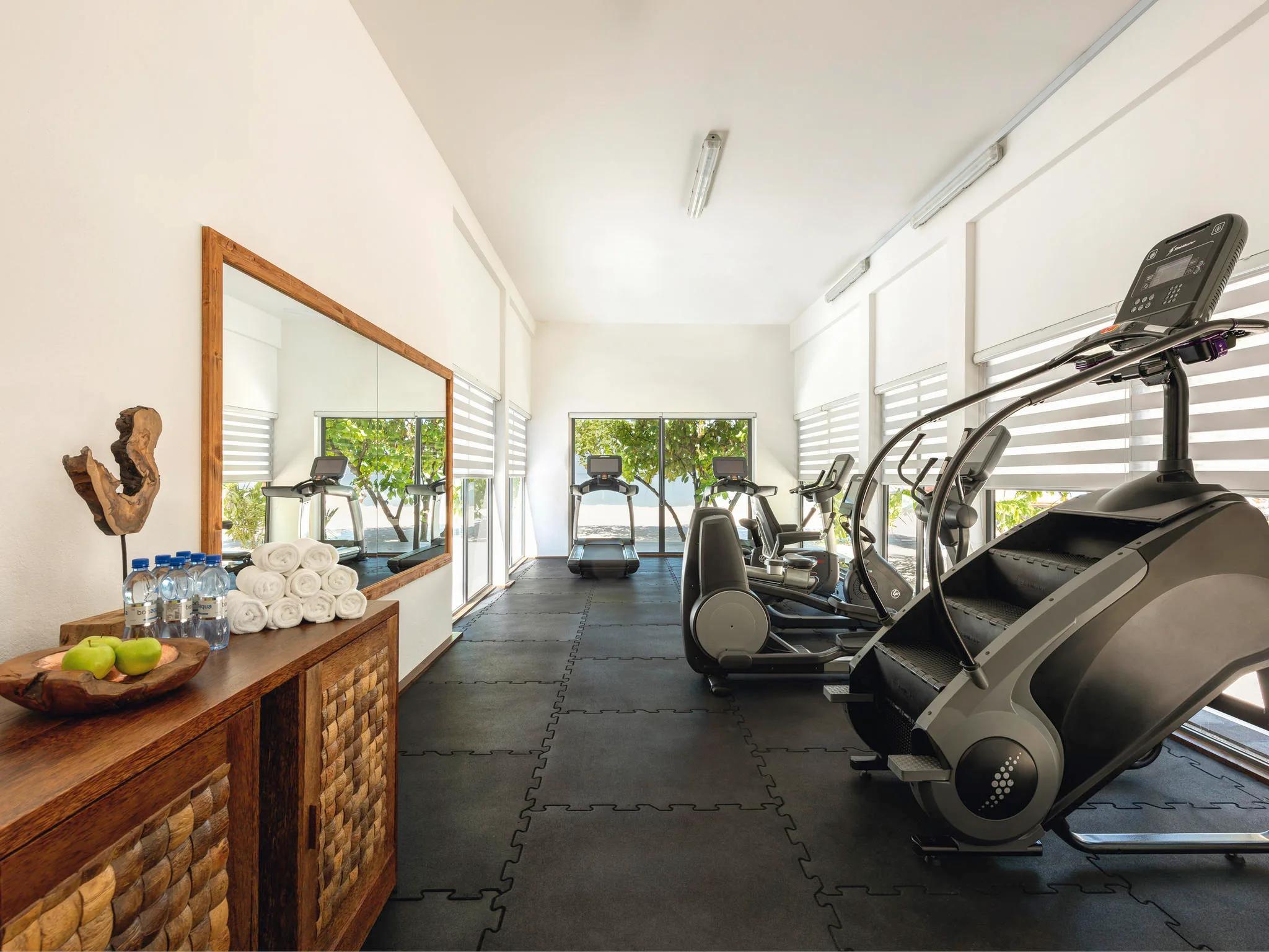 An open, mirror-lined fitness centre with a row of treadmills and cross-trainers facing floor-to-ceiling windows onto greenery