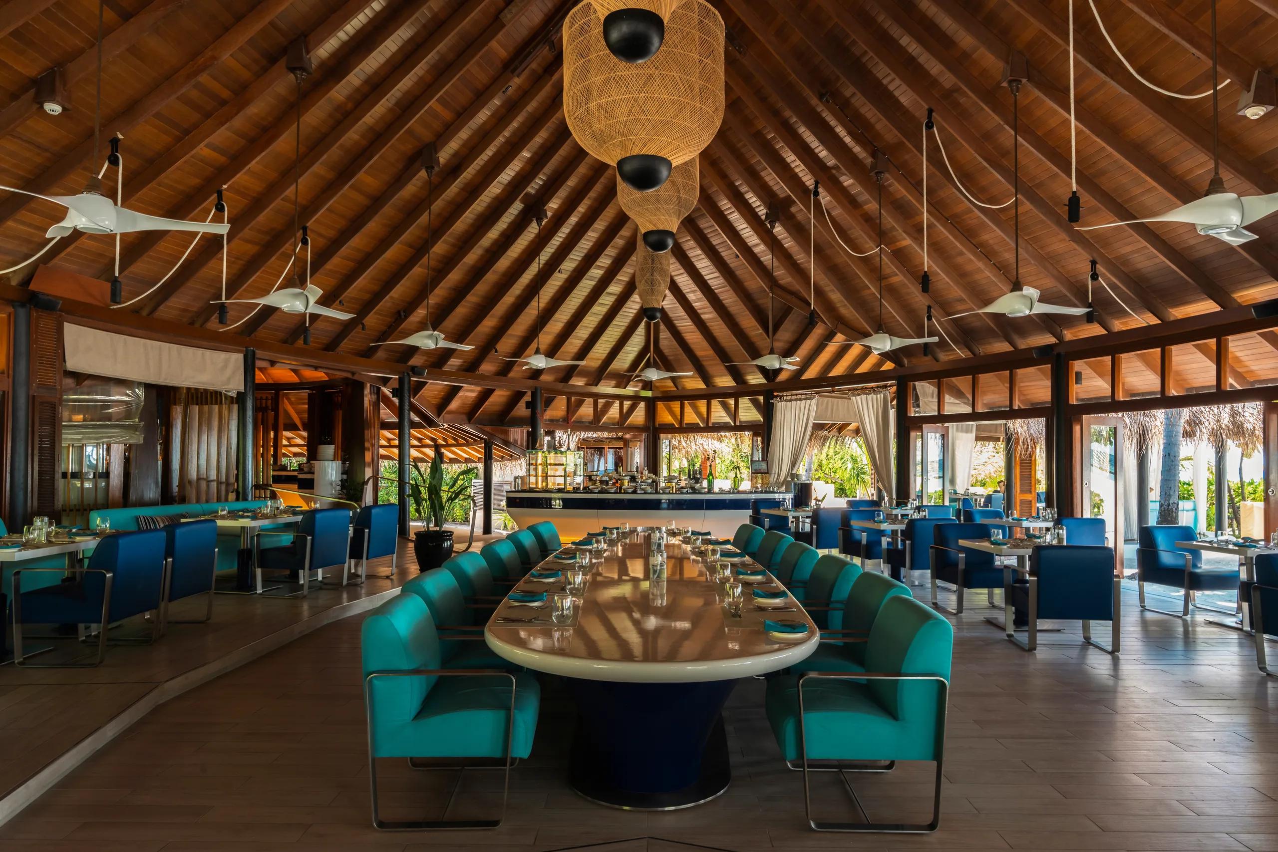 The interior of Jahaz, Constance Halaveli's main restaurant, showing its high thatched roof, ceiling fans, and a long central table set with blue and teal upholstered chairs