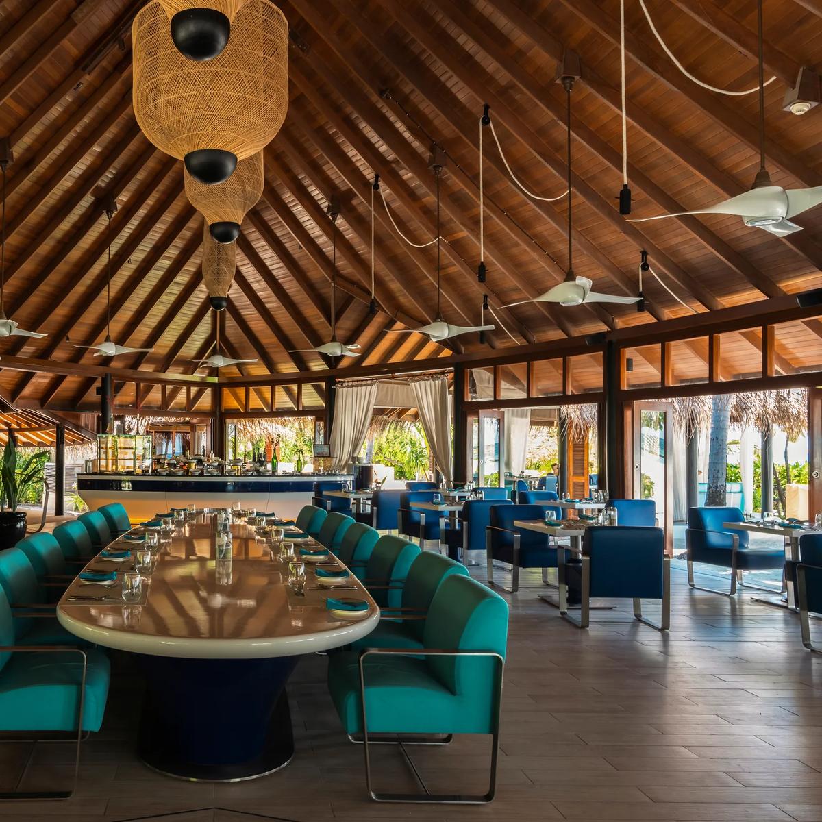 The interior of Jahaz, Constance Halaveli's main restaurant, showing its high thatched roof, ceiling fans, and a long central table set with blue and teal upholstered chairs