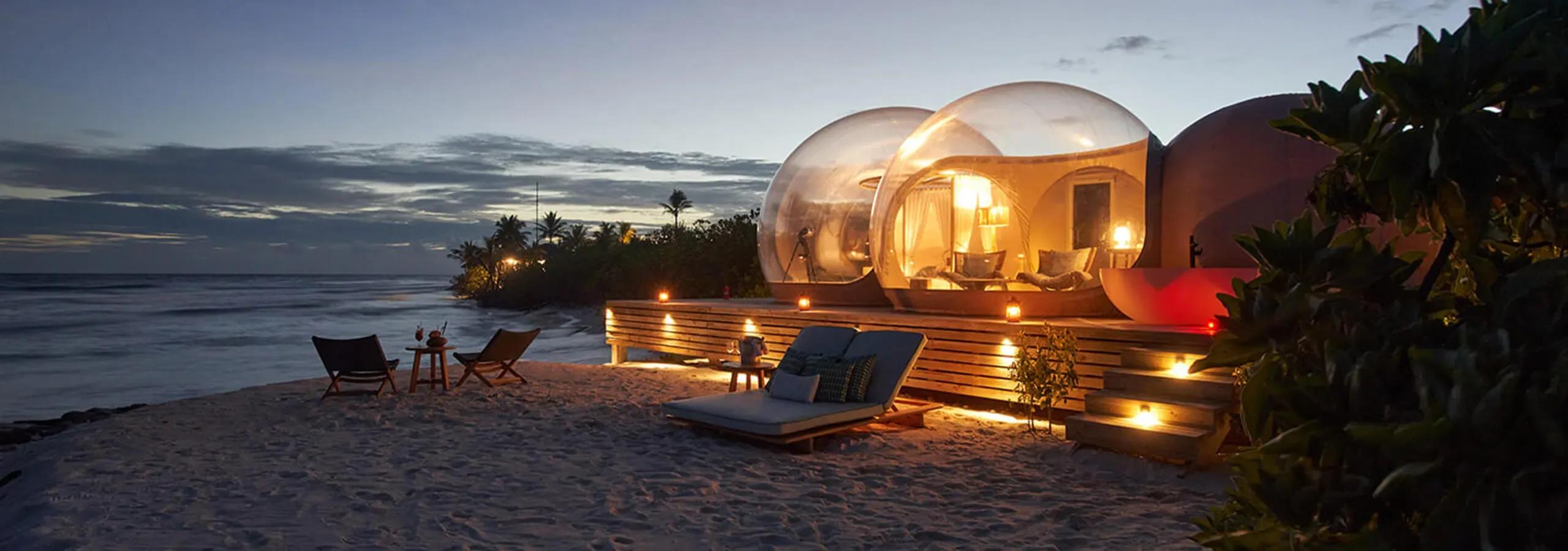 Illuminated glass-domed Beach Bubble Tents on the sand at dusk, with loungers and lit lanterns set out on the beach in front