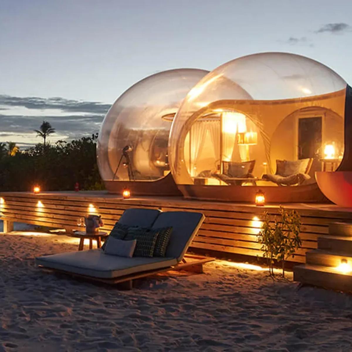 Illuminated glass-domed Beach Bubble Tents on the sand at dusk, with loungers and lit lanterns set out on the beach in front