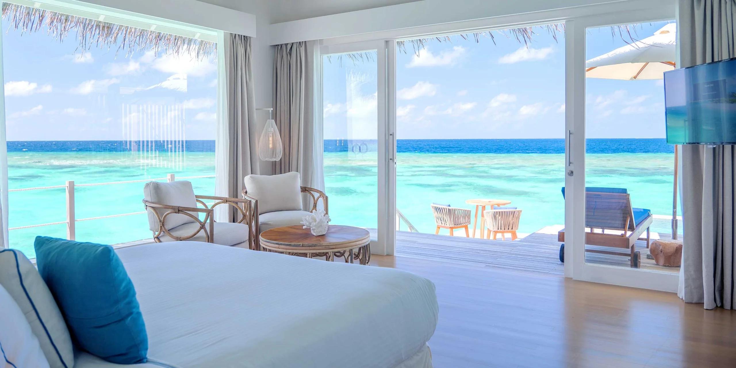 An overwater Water Villa with Pool's lounge area, rattan chairs facing sliding doors onto the deck and open lagoon