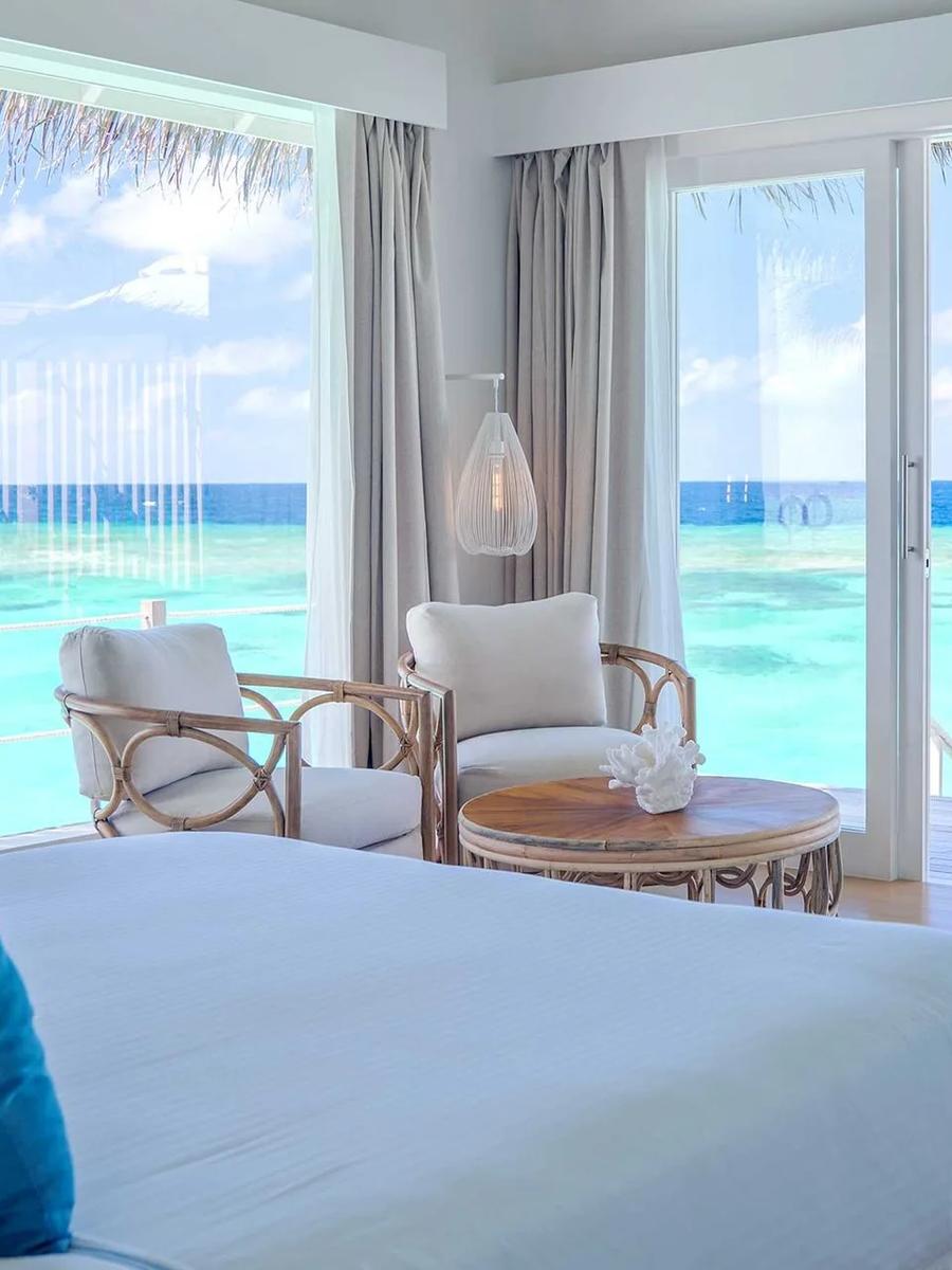 An overwater Water Villa with Pool's lounge area, rattan chairs facing sliding doors onto the deck and open lagoon