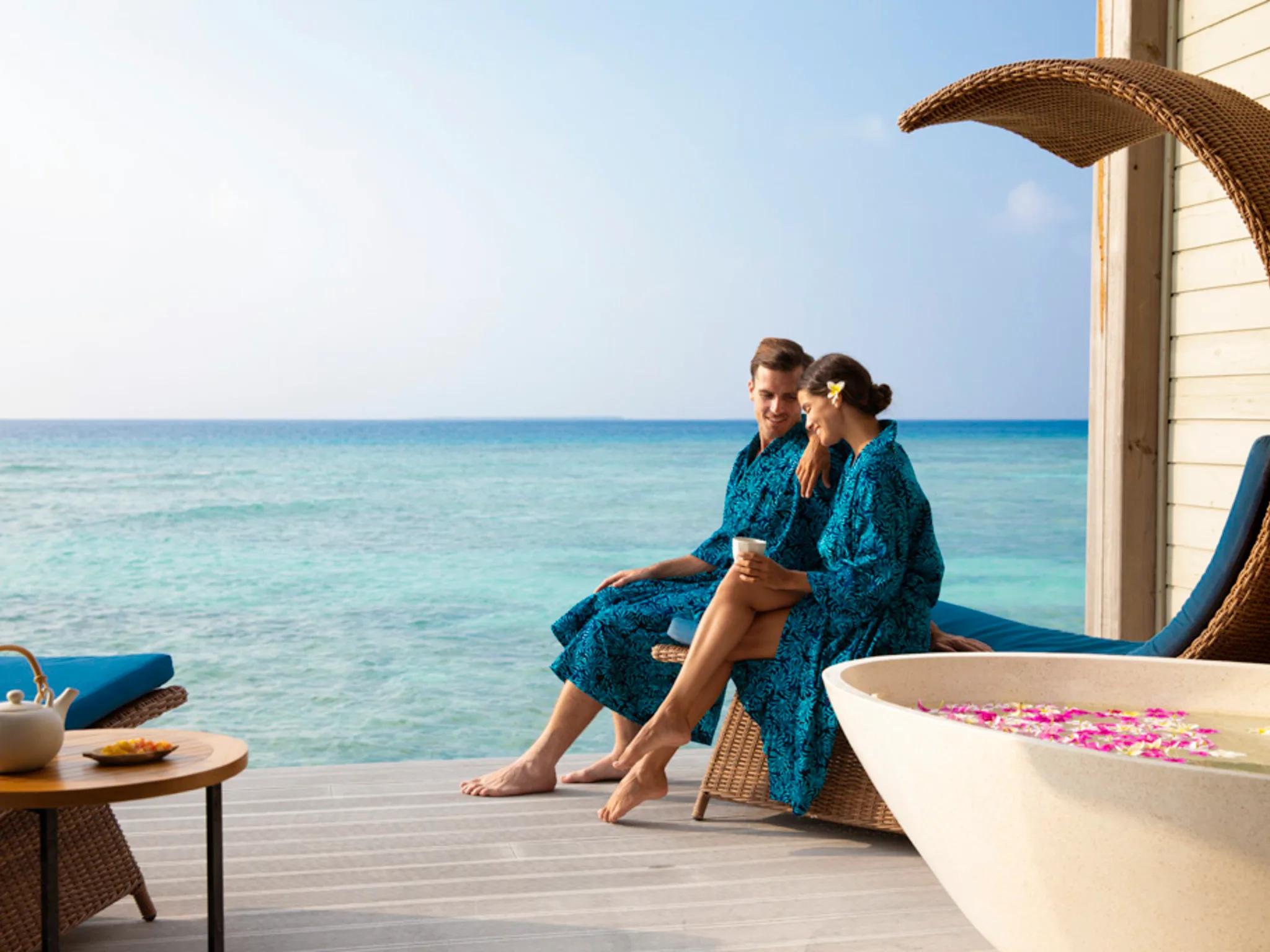A couple in blue robes sitting together on an overwater deck at golden hour, a stone bathtub filled with flower petals beside them
