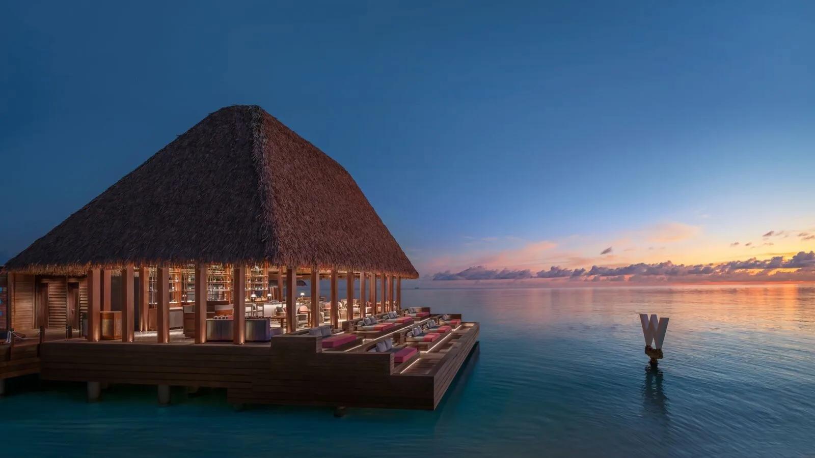 SIP's overwater pavilion at W Maldives at dusk, with the resort's W-shaped marker standing in the lagoon beyond