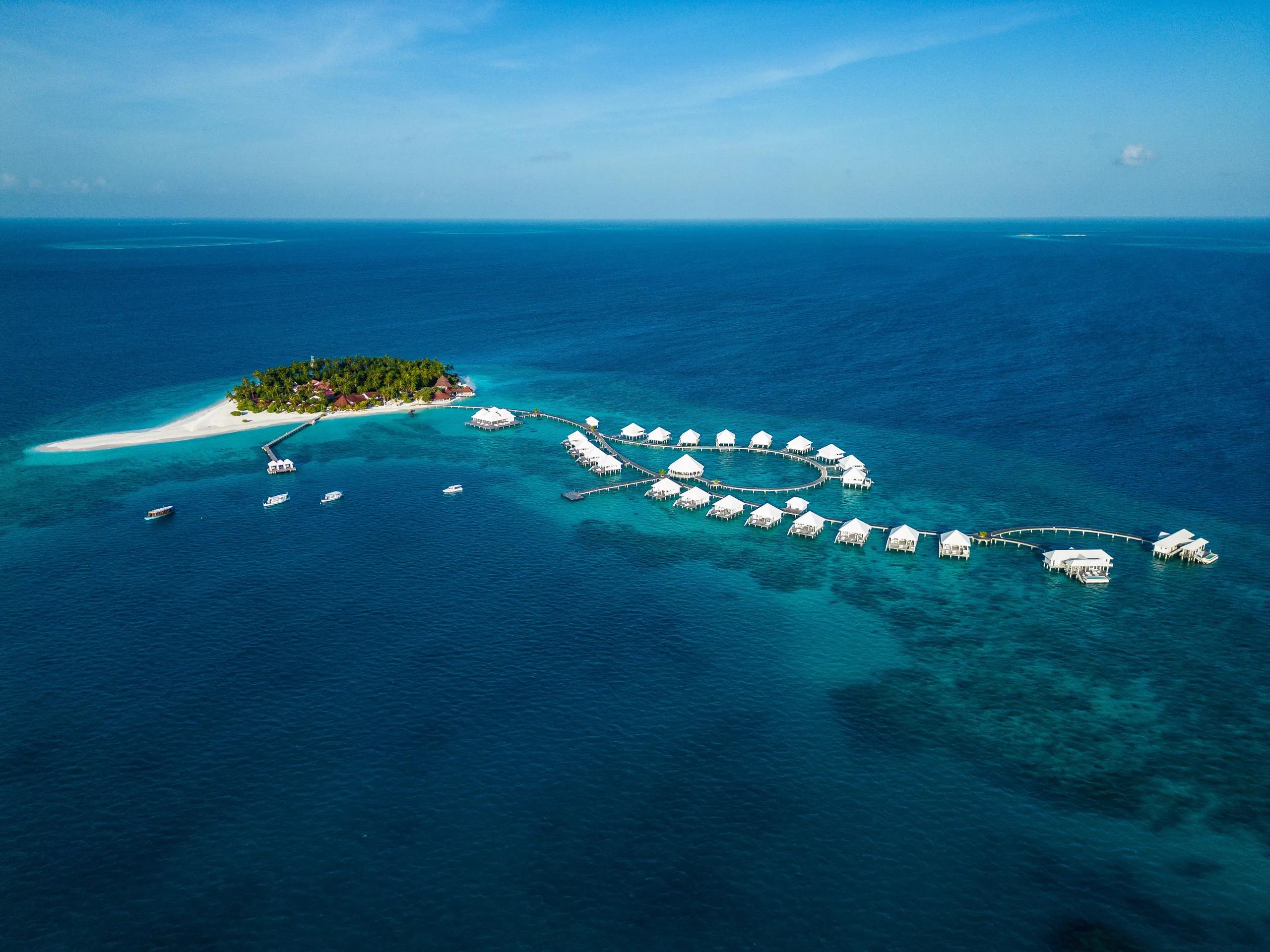 Aerial view of Diamonds Thudufushi's palm-covered island and its curving overwater villa jetty reaching out toward the reef edge, South Ari Atoll