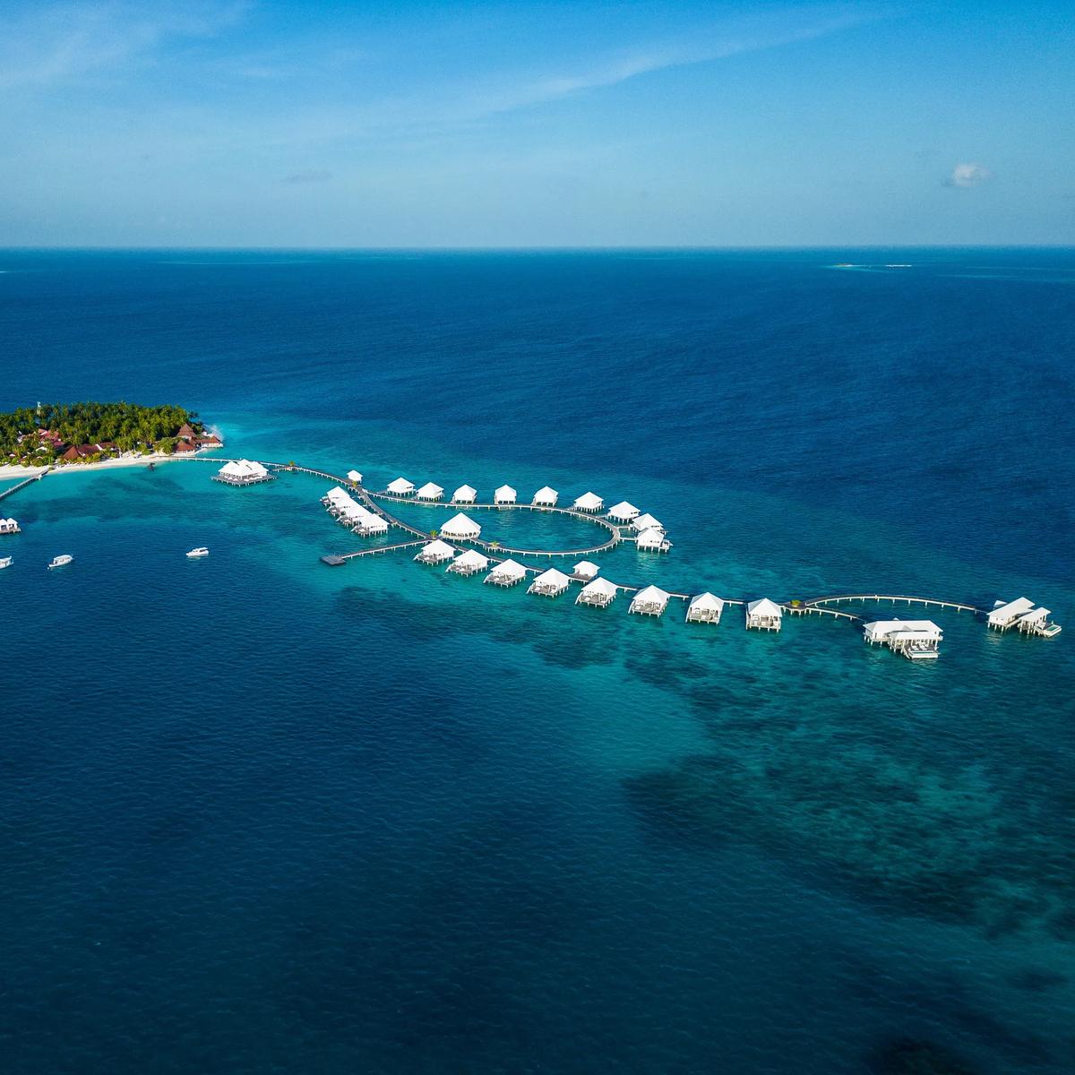 Aerial view of Diamonds Thudufushi's palm-covered island and its curving overwater villa jetty reaching out toward the reef edge, South Ari Atoll