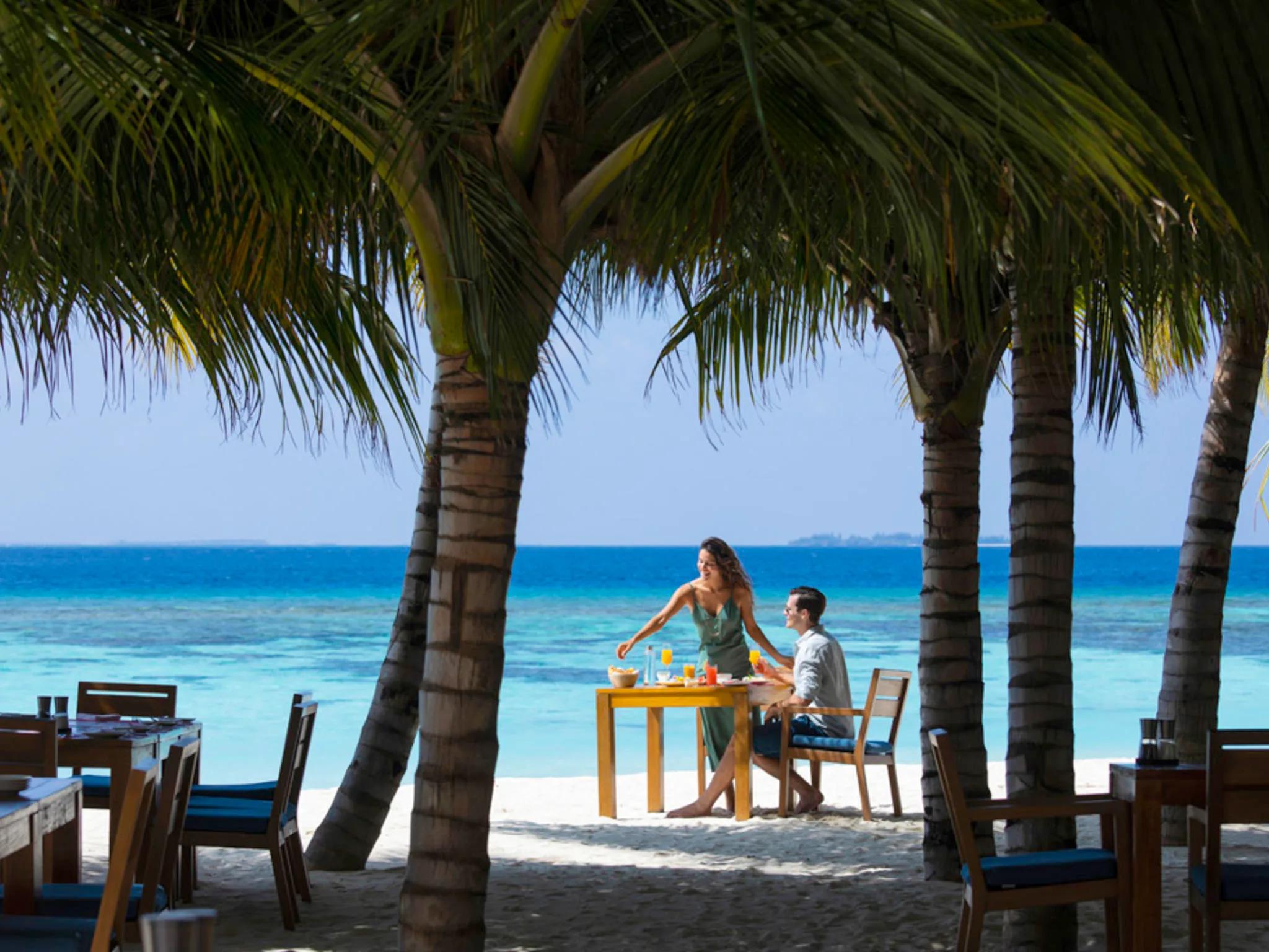 A couple seated at a small beach table shaded by palm trees, fresh juices and a light breakfast spread between them, turquoise lagoon beyond
