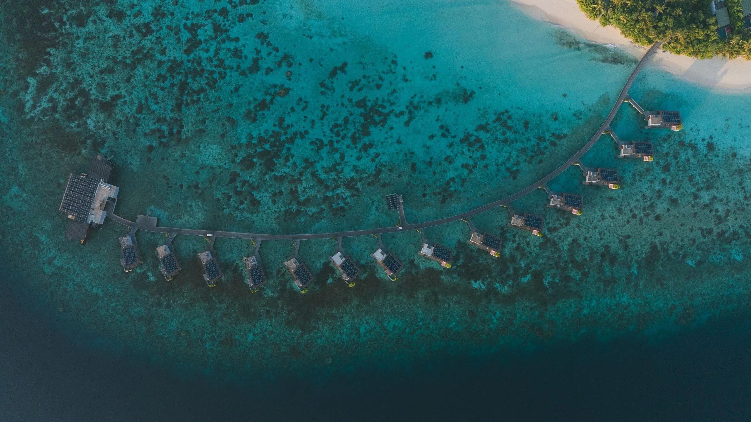 Aerial view of Park Hyatt Maldives Hadahaa's overwater villas curving from the main jetty toward the beach, the reef visible beneath the clear lagoon
