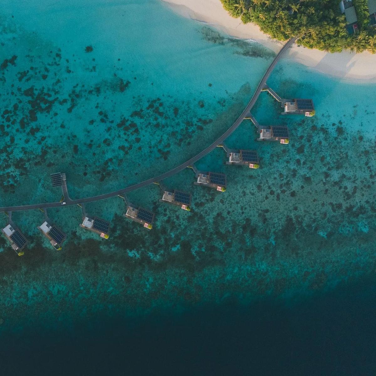 Aerial view of Park Hyatt Maldives Hadahaa's overwater villas curving from the main jetty toward the beach, the reef visible beneath the clear lagoon