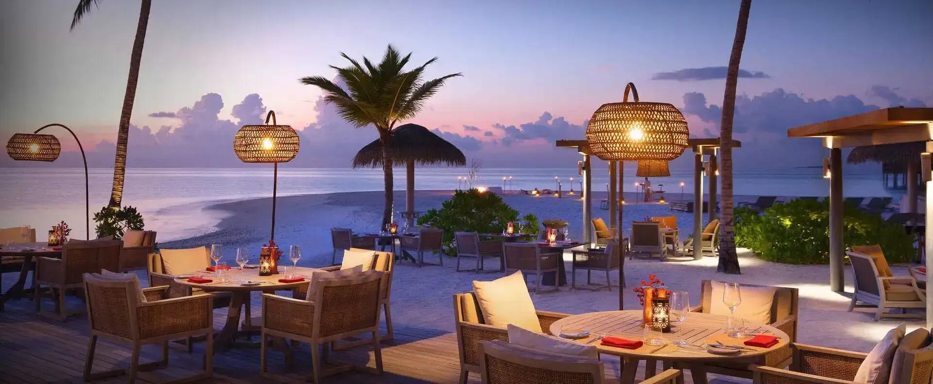 Vakku's beachfront dining tables set for dinner among palm trees at dusk, rattan pendant lamps lit against a fading sky
