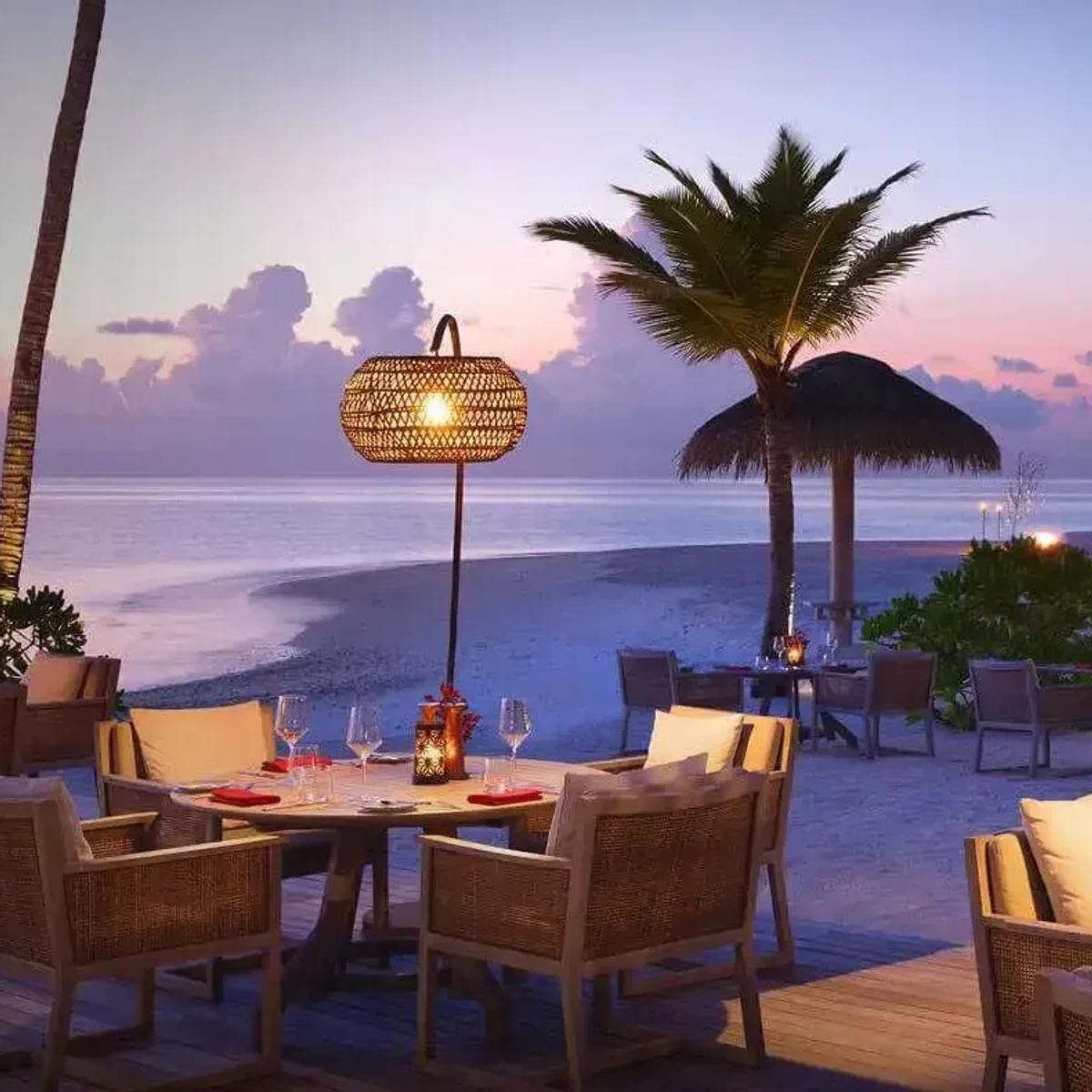 Vakku's beachfront dining tables set for dinner among palm trees at dusk, rattan pendant lamps lit against a fading sky