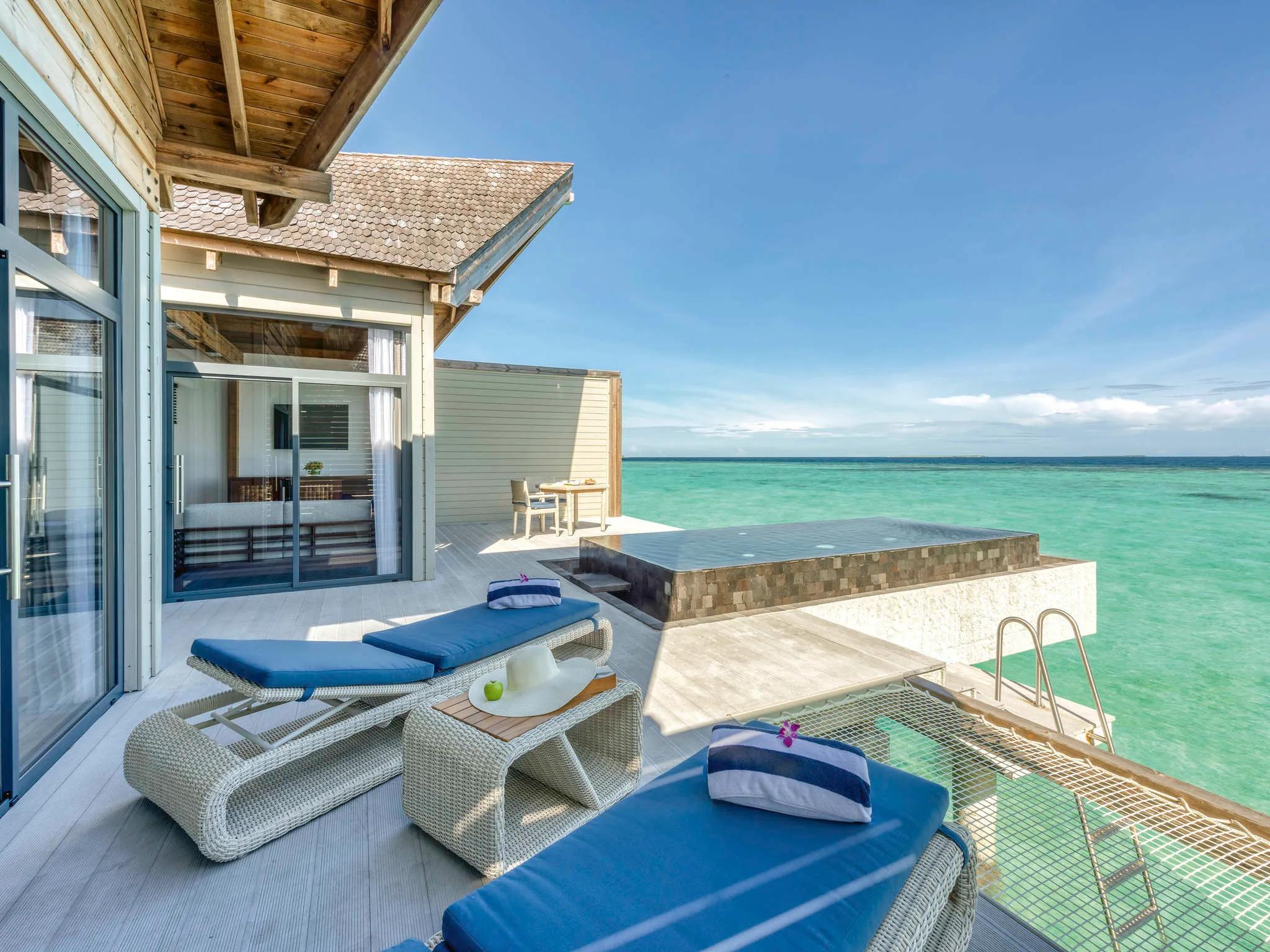 Deck of an overwater pool villa with a private pool, two blue sun loungers and a sun hat, open ocean stretching to the horizon