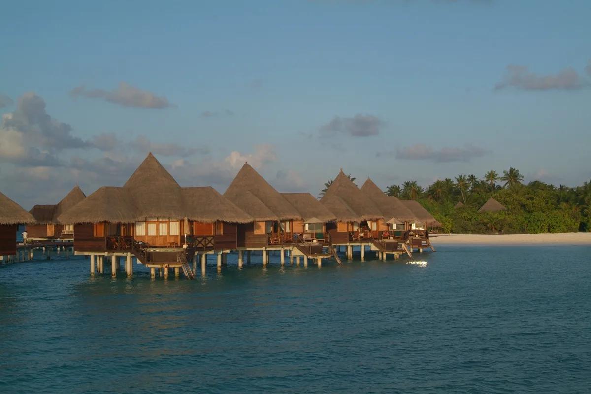 A row of overwater villas on stilts above the lagoon at dusk, beach and palm trees behind