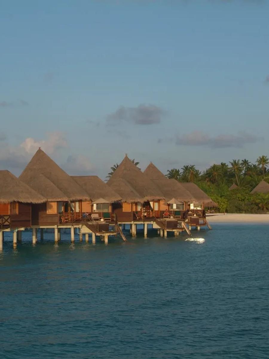 A row of overwater villas on stilts above the lagoon at dusk, beach and palm trees behind