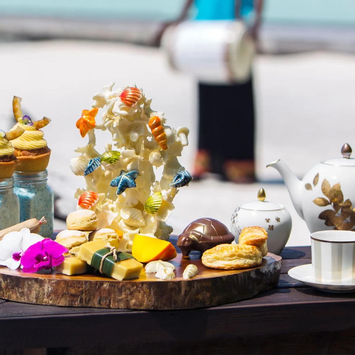 A High Tea spread on the beach — scones, pastries, a coral-shaped sugar sculpture and a pot of tea — a Boduberu drummer performing in the background