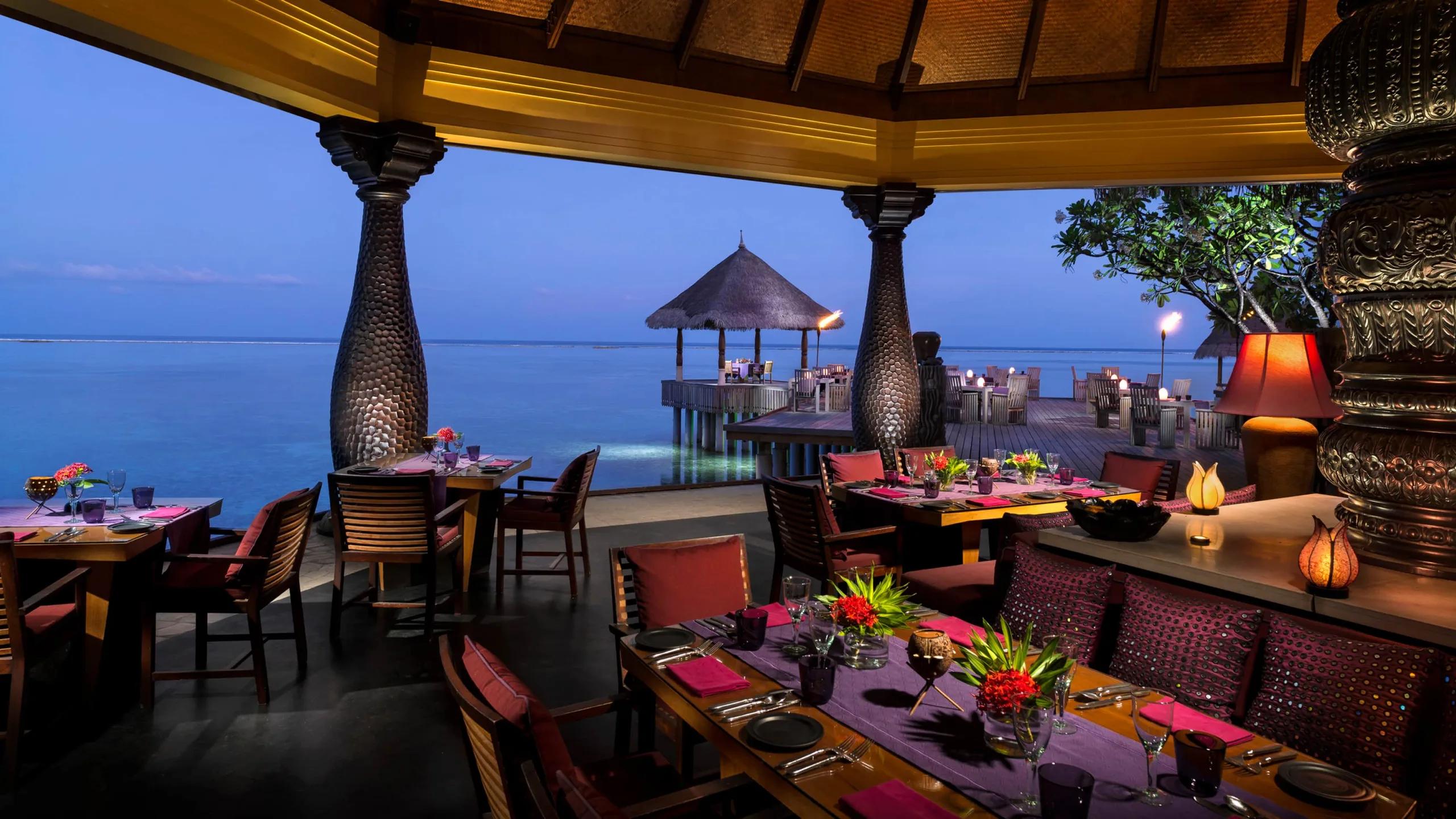 Baraabaru's overwater dining terrace at dusk, tables set beneath a thatched roof beside the lagoon