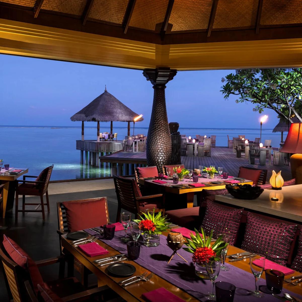 Baraabaru's overwater dining terrace at dusk, tables set beneath a thatched roof beside the lagoon