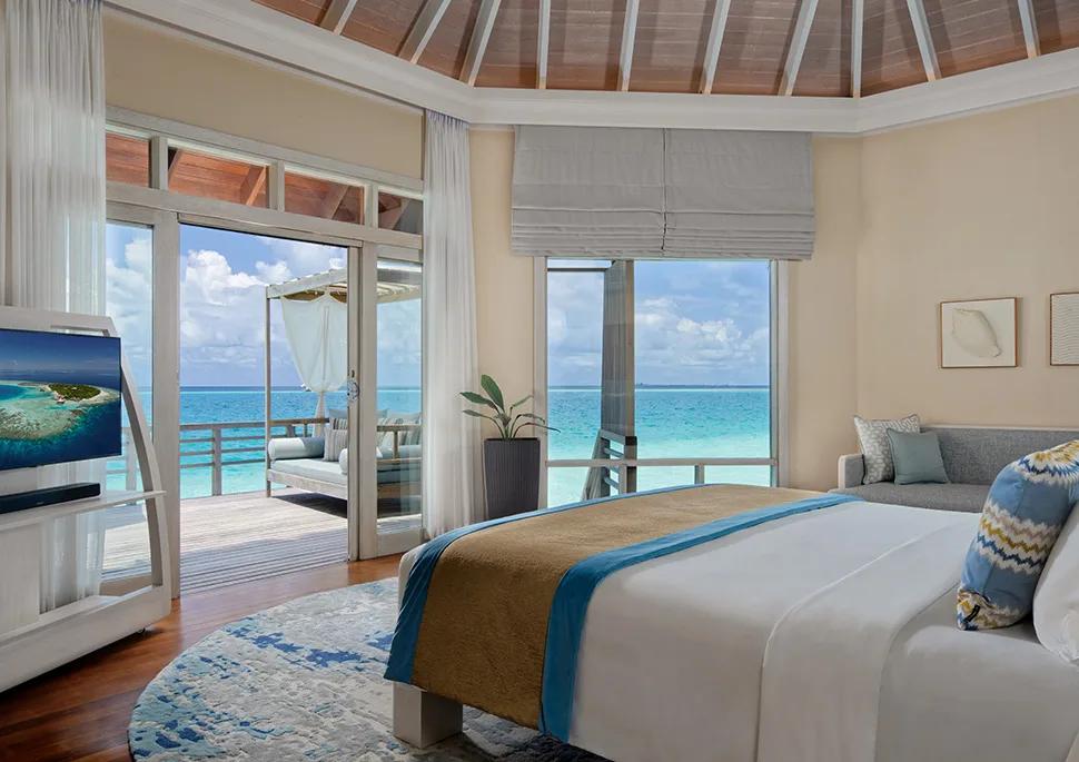 A Water Villa bedroom with sliding doors open to a private overwater deck and daybed, the lagoon visible on every side