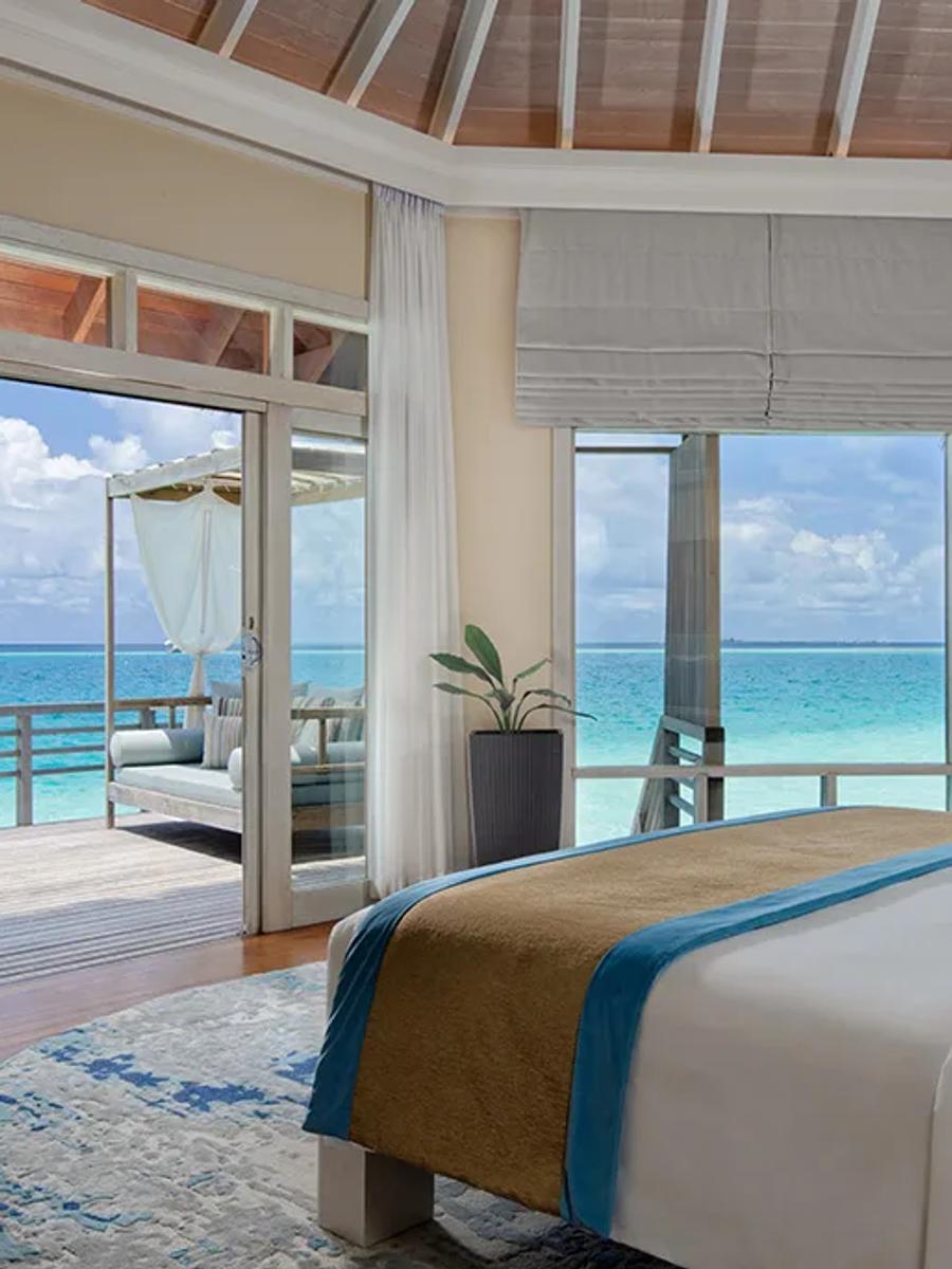 A Water Villa bedroom with sliding doors open to a private overwater deck and daybed, the lagoon visible on every side