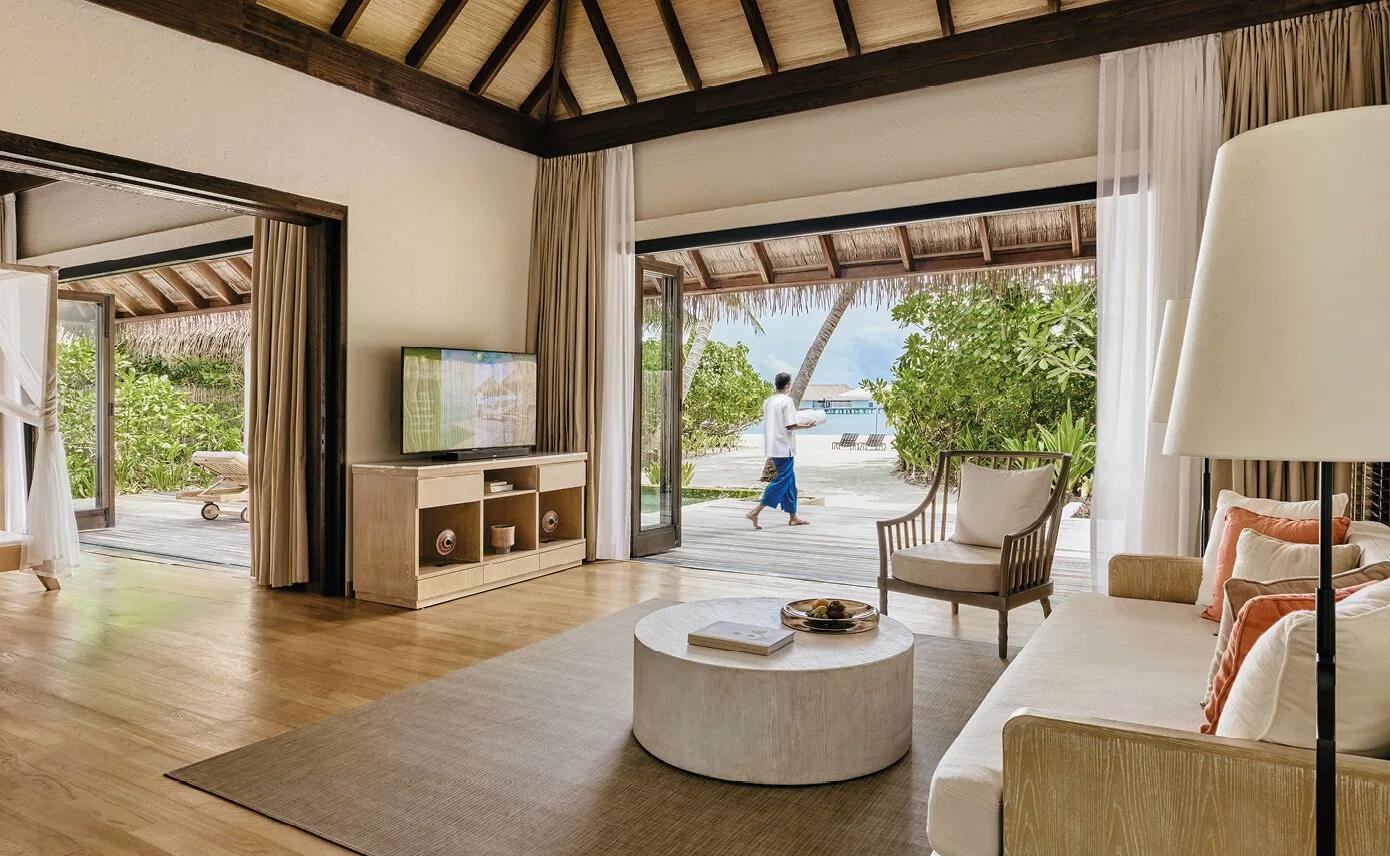 Open-plan villa living room with sliding doors onto a wooden deck, a staff member crossing the lawn toward the beach and overwater villas in the distance