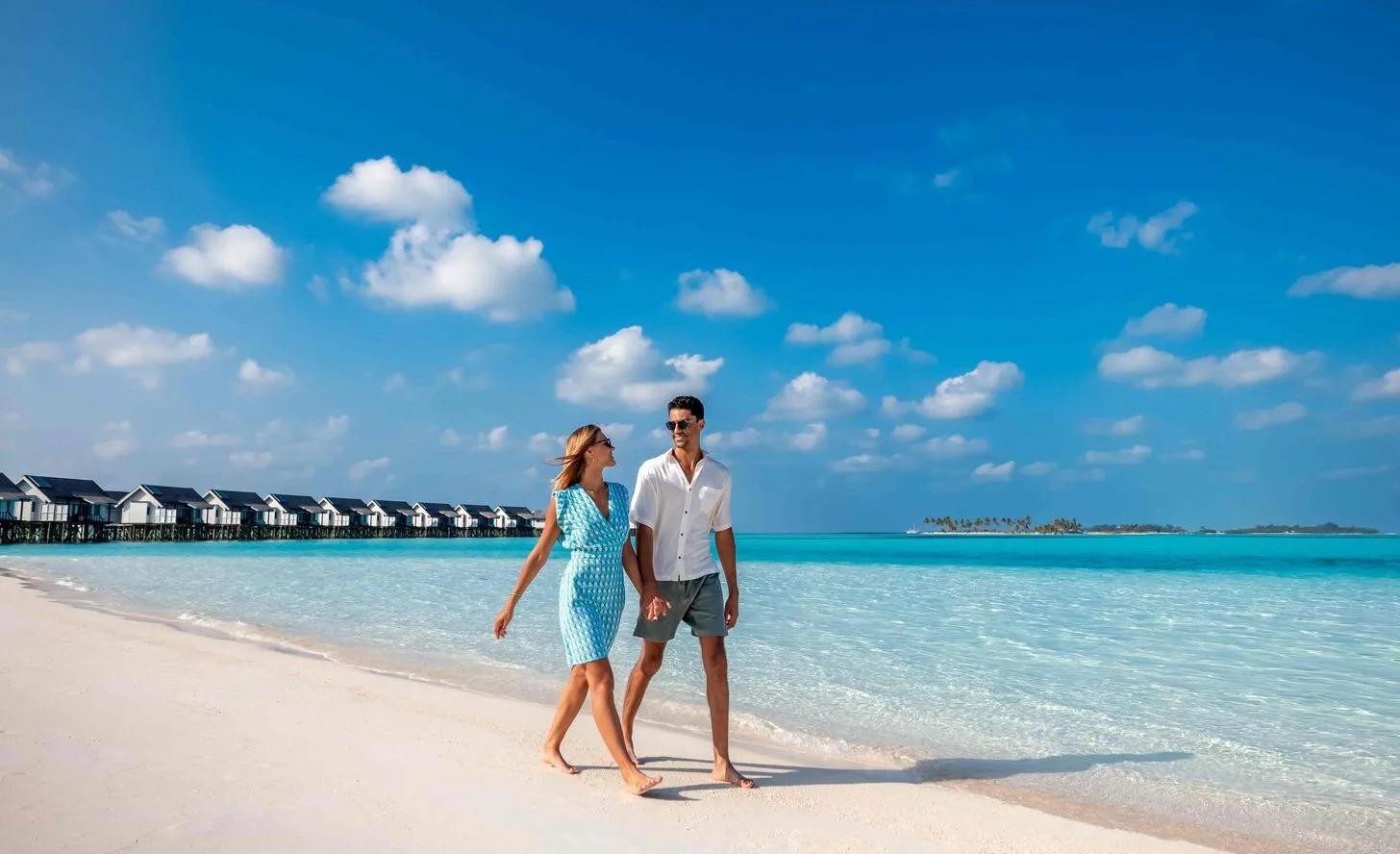 A couple walking the beach at low tide, overwater villas and a neighbouring island behind them
