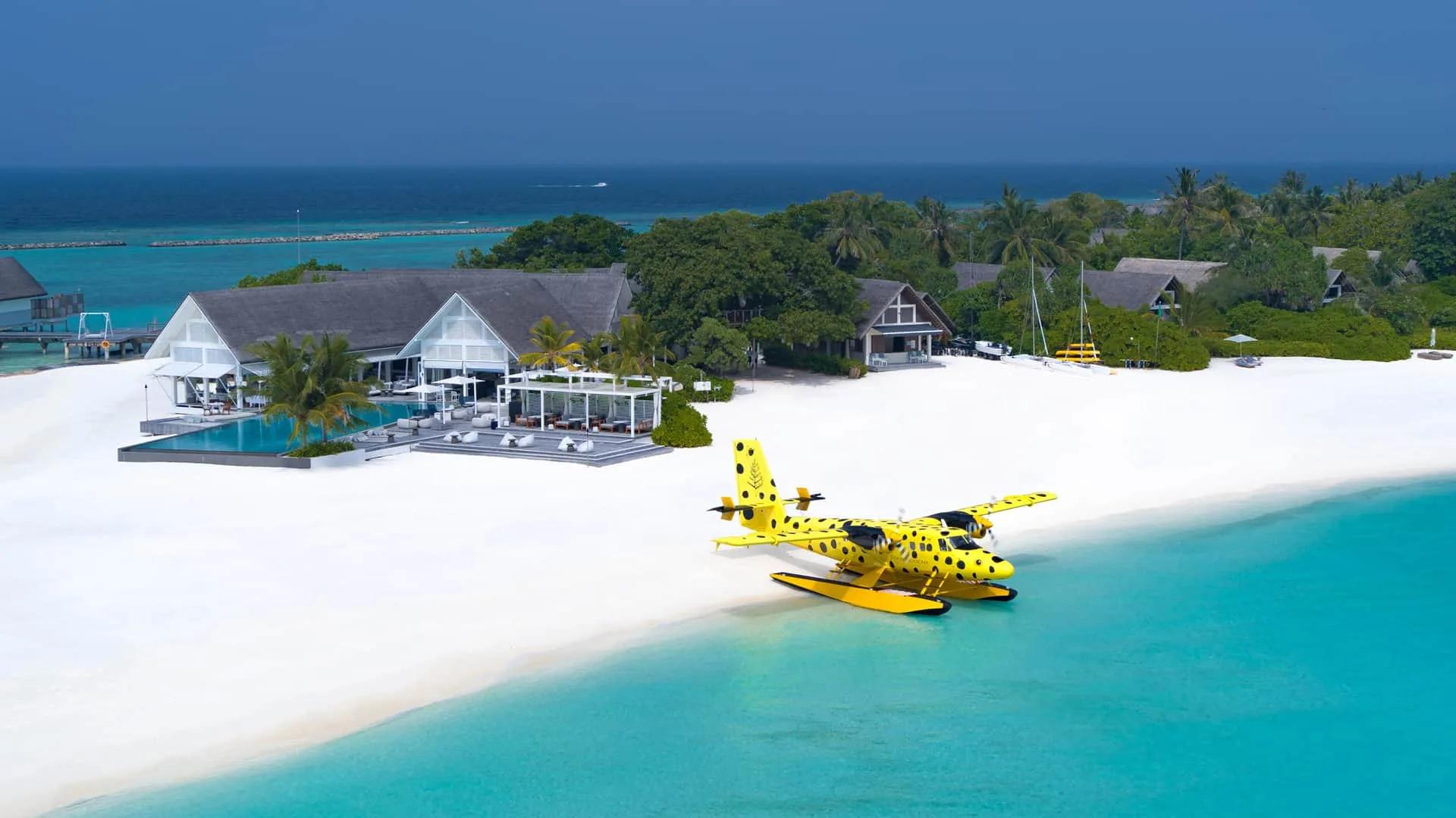 Aerial view of Four Seasons Landaa Giraavaru's beach club and jetty, with a spotted yellow seaplane on the shallows and overwater villas along the reef edge