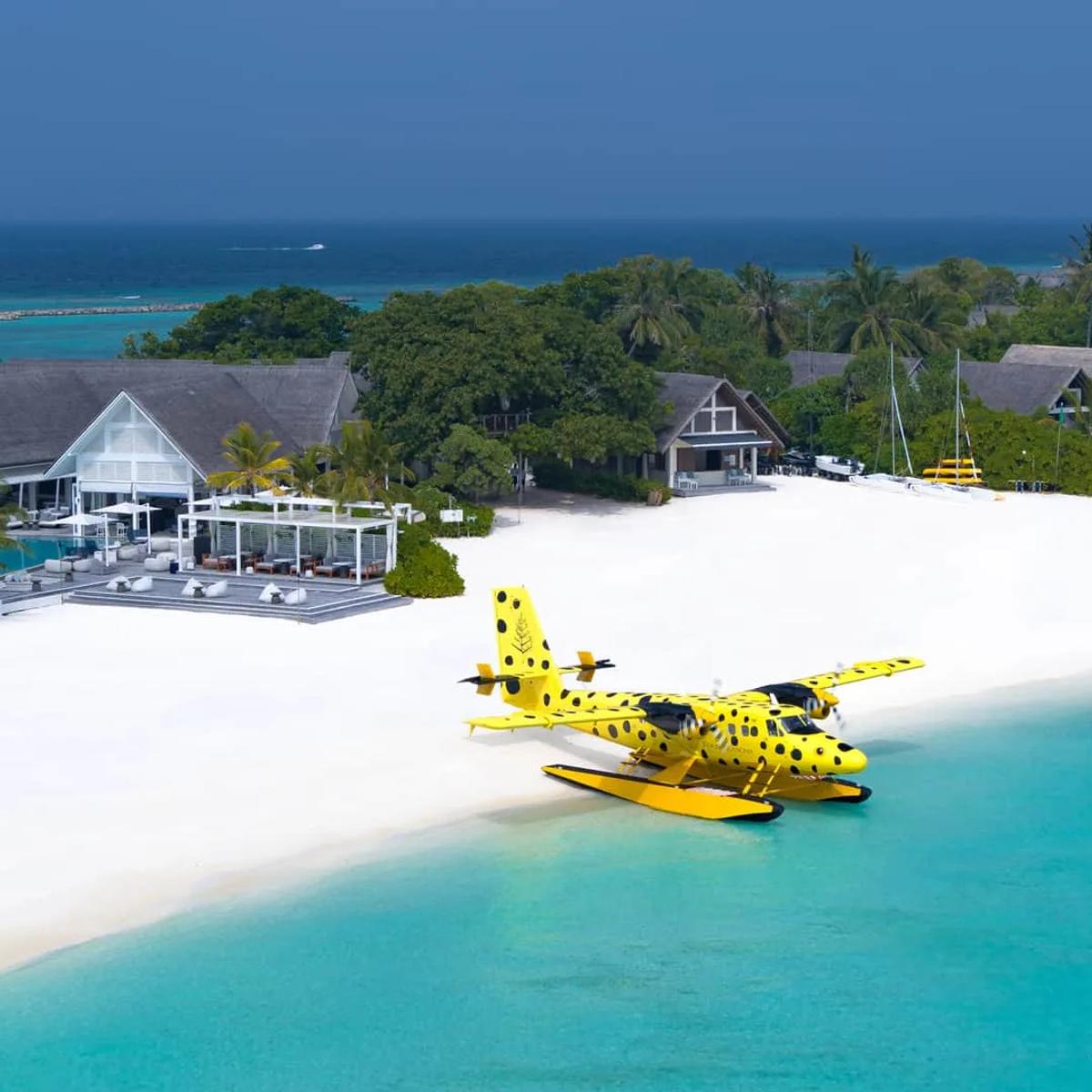 Aerial view of Four Seasons Landaa Giraavaru's beach club and jetty, with a spotted yellow seaplane on the shallows and overwater villas along the reef edge