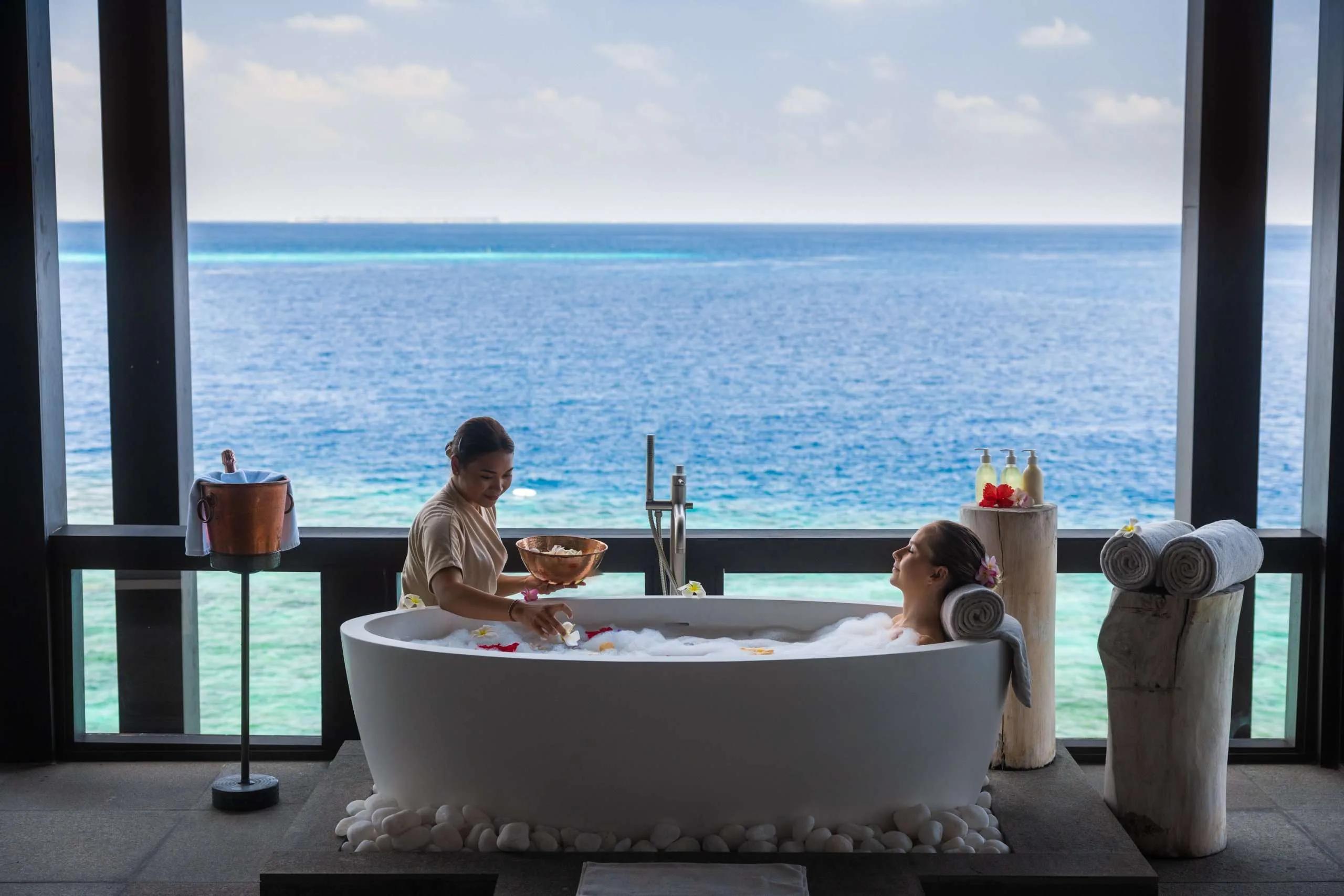 A therapist scattering flower petals into an overwater bathtub for a reclining guest, lagoon behind