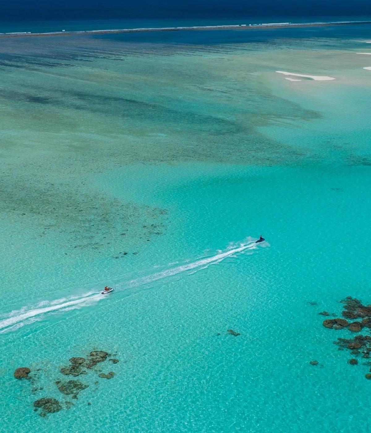 Aerial view of a jet ski crossing the turquoise lagoon at SO/ Maldives, its wake cutting across the shallow reef flat