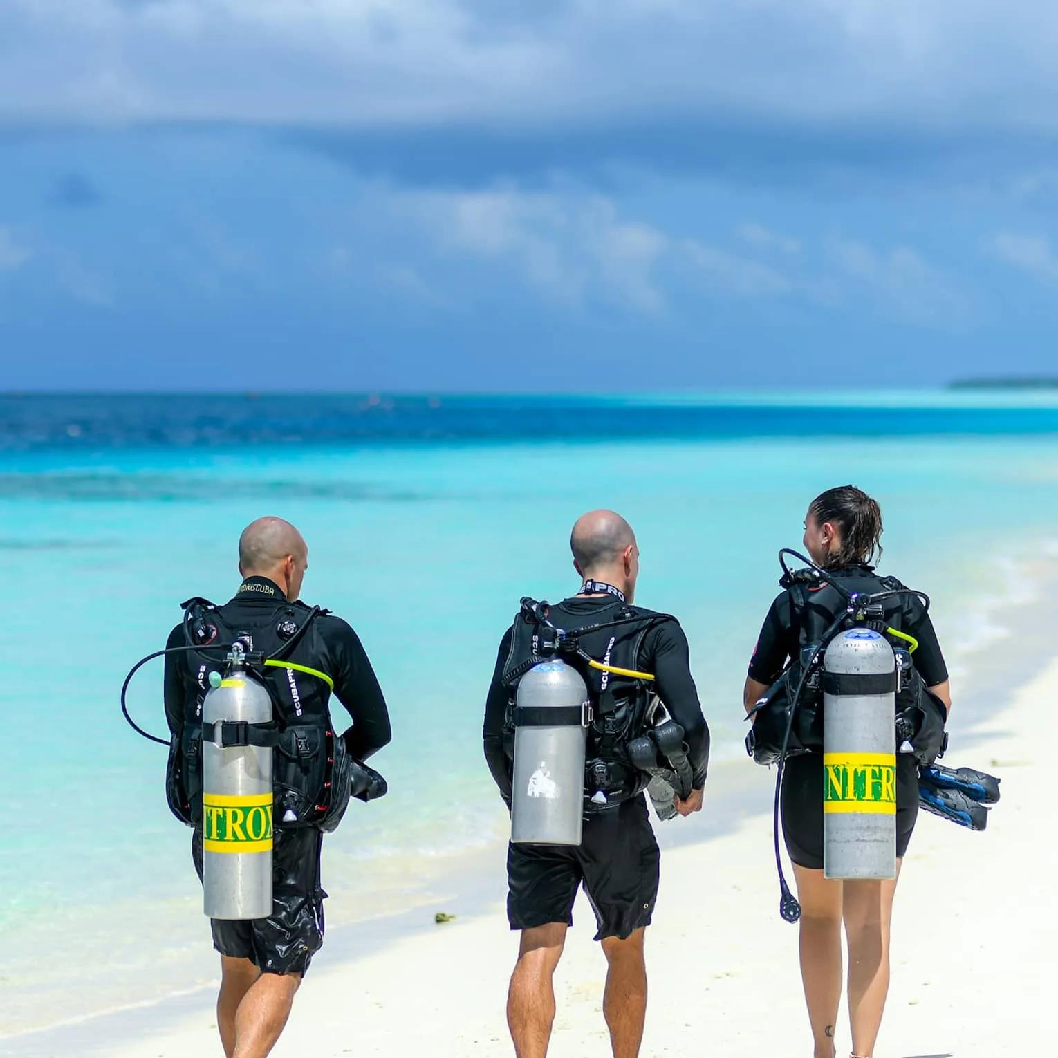 Three divers walking to the water with Nitrox tanks, the resort's beach behind them